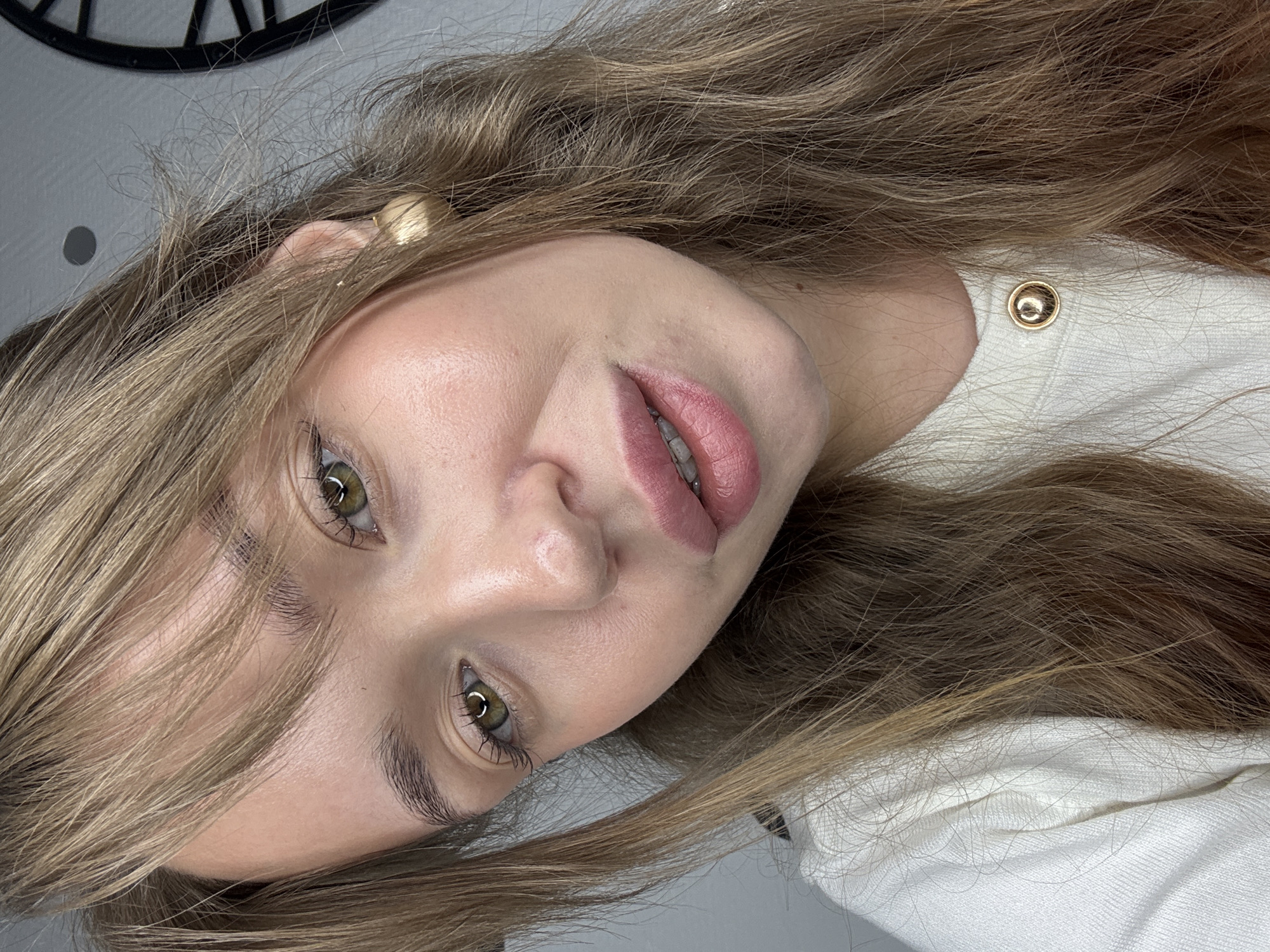 A close-up portrait features a young woman with long, light brown hair, which appears slightly tousled and falls around her face and shoulders. The image is rotated clockwise, showing her head tilted to the right. She looks directly at the camera with her green or hazel eyes. Her lips are slightly parted, revealing a glimpse of her teeth, and are adorned with a pinkish lip color. She has well-defined eyebrows and appears to be wearing mascara.
She has a small gold stud earring visible in her left ear. She is wearing a white garment, possibly a sweater or top, with a textured knit. A prominent gold button is visible on the shoulder of her garment. The background is a plain light gray wall, with a partial view of a dark, spoked circular object, possibly a clock or decor, in the upper left corner of the original image. No specific activity beyond posing for the camera is discernible. There is no visible text in the image. The general setting is an indoor space, located in Chaikovsky, Russia, as per the provided context.