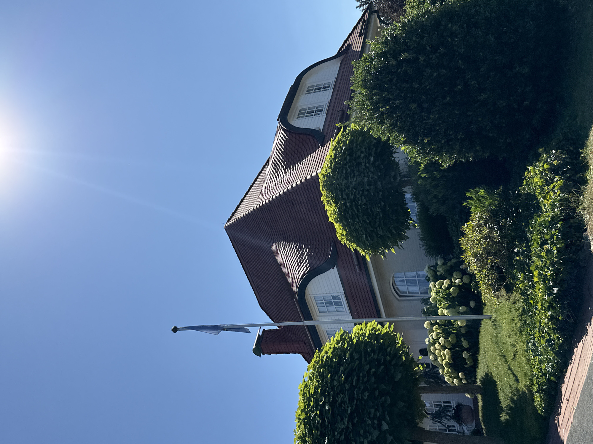 The image depicts a two-story house with a distinctive mansard roof covered in red tiles, situated in Bad Lauterberg im Harz, Germany. The house has white siding and several windows, including dormer windows on the upper floor. A prominent brick chimney is visible on the left side of the roof. In front of the house, there is a well-maintained garden with manicured trees, bushes, and a large hydrangea bush in bloom. A flagpole with a blue flag is planted in the lawn to the left of the house. The sky is clear blue, and the sun is shining brightly, casting some glare on the upper part of the image. The foreground shows a brick pathway. The overall scene conveys a sense of a well-kept residential property on a sunny day.