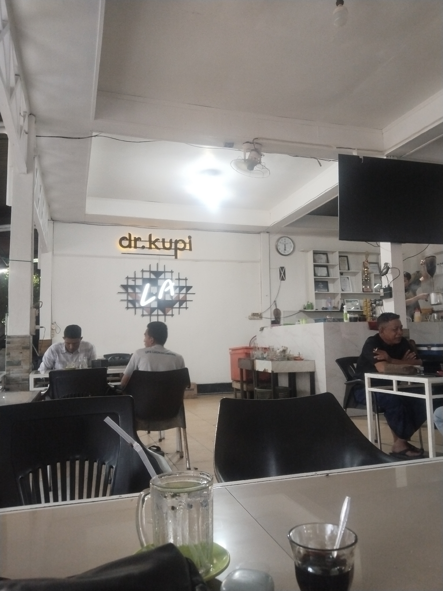 The image depicts the interior of "dr.kupi," a cafe located in Lhokseumawe, Indonesia.
Three men are visible in the scene. Two men are seated at a table in the foreground, while a third man is seated at a table in the background. The table in the foreground has two glasses on it, one with a straw.
The cafe's interior has white walls. A clock is visible on the wall behind the counter.