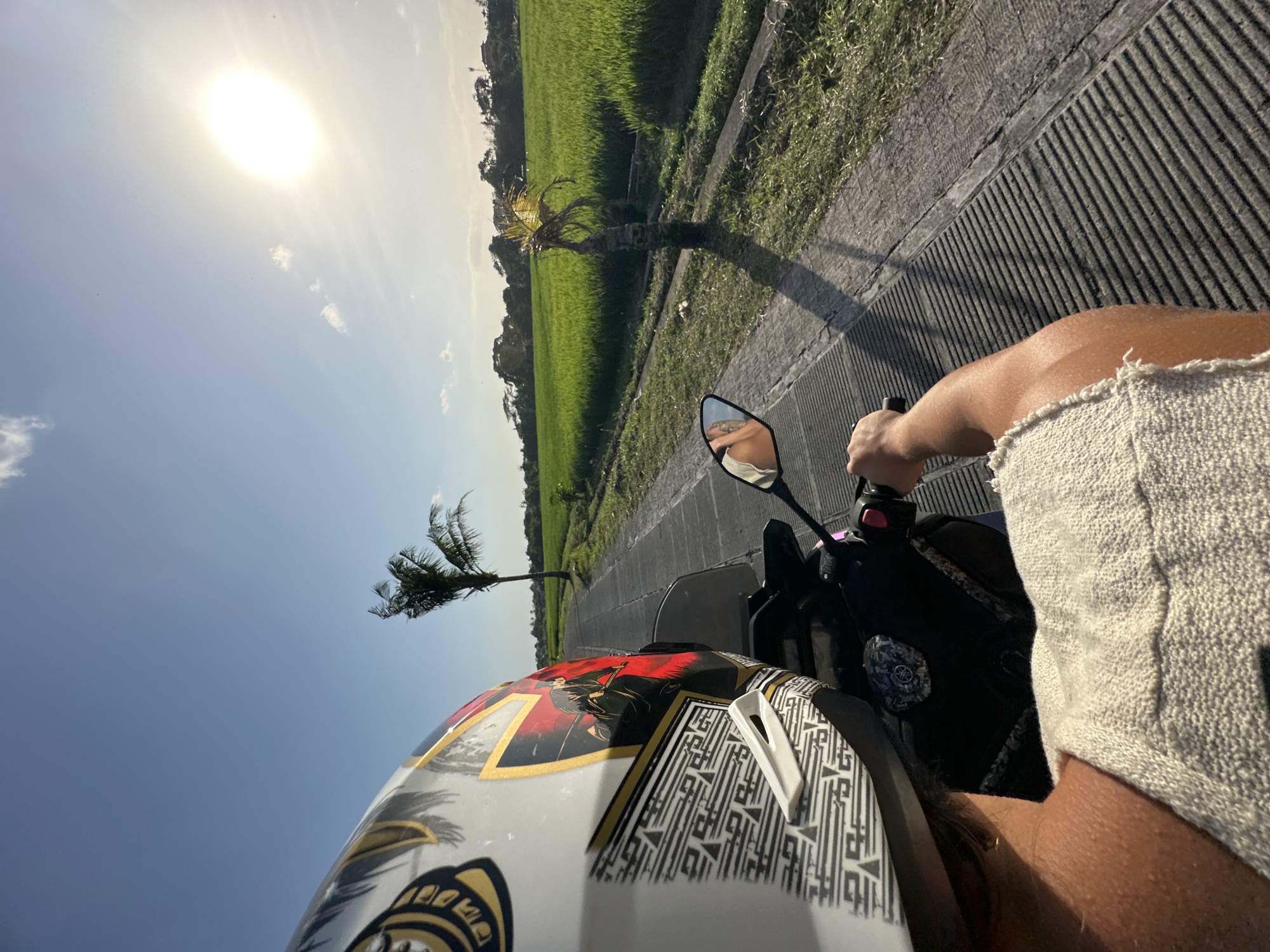 A first-person perspective shot shows someone riding a scooter on a paved road through a rural area, likely in North Kuta, Indonesia. The rider, wearing a helmet with a graphic design of a samurai warrior, has their right hand on the handlebar. The road curves to the right, bordered by a lush green rice paddy on the right and a thin strip of grass on the left. In the background, a single palm tree stands tall, and beyond that, more greenery and some houses are visible under a bright, sunny sky with scattered clouds. The sun is high and bright, casting long shadows. A side mirror on the scooter reflects a glimpse of the rider's face and the sky.