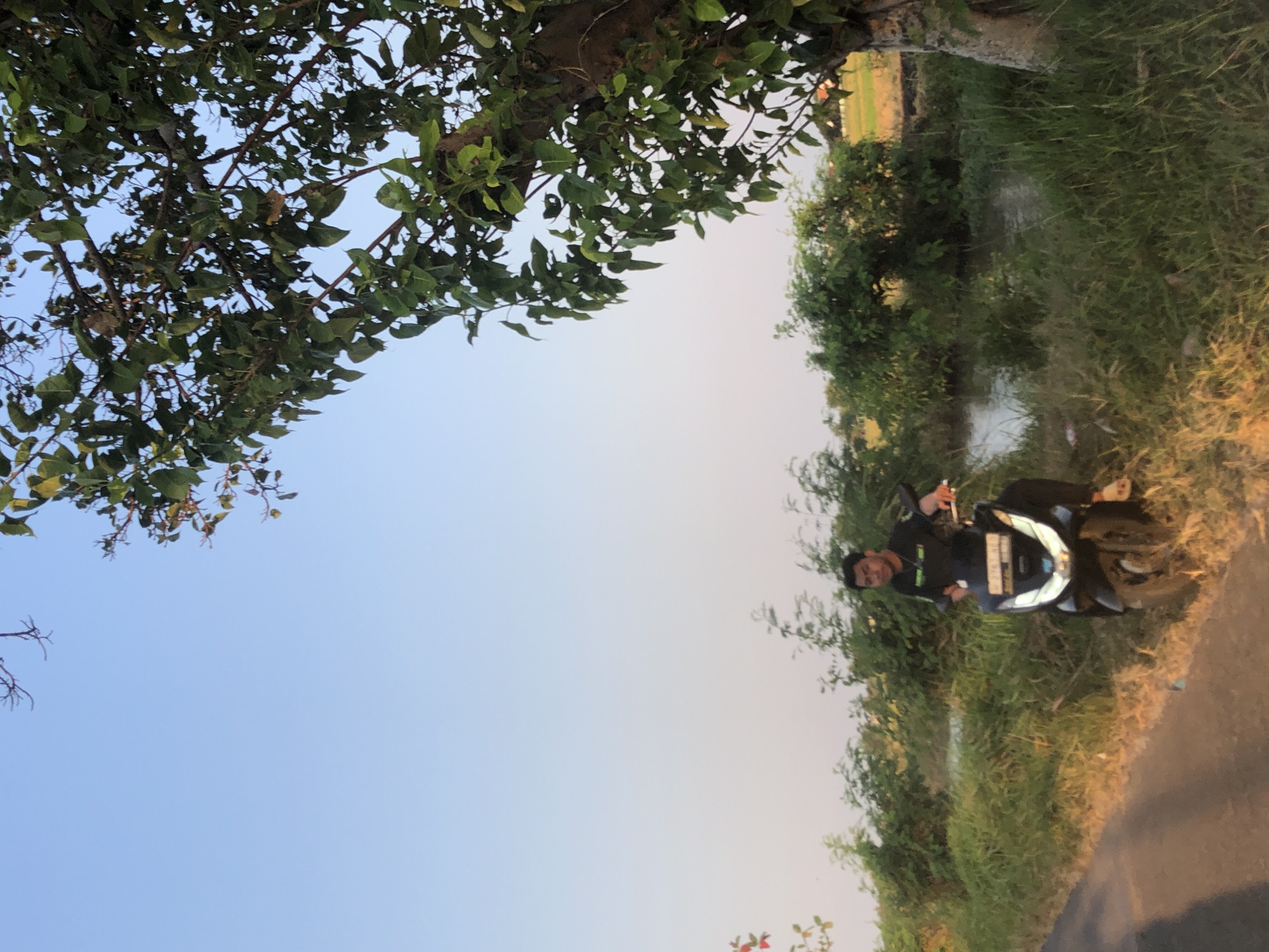 In Keboguyang, Indonesia, a young man is seated on a black motorcycle parked on the side of a dirt road. He is wearing a black t-shirt and dark pants. The motorcycle's headlight is illuminated. The road is bordered by tall green grass and a small body of water. In the background, lush green foliage and trees are visible under a clear blue sky. The overall scene depicts a rural landscape during daylight.