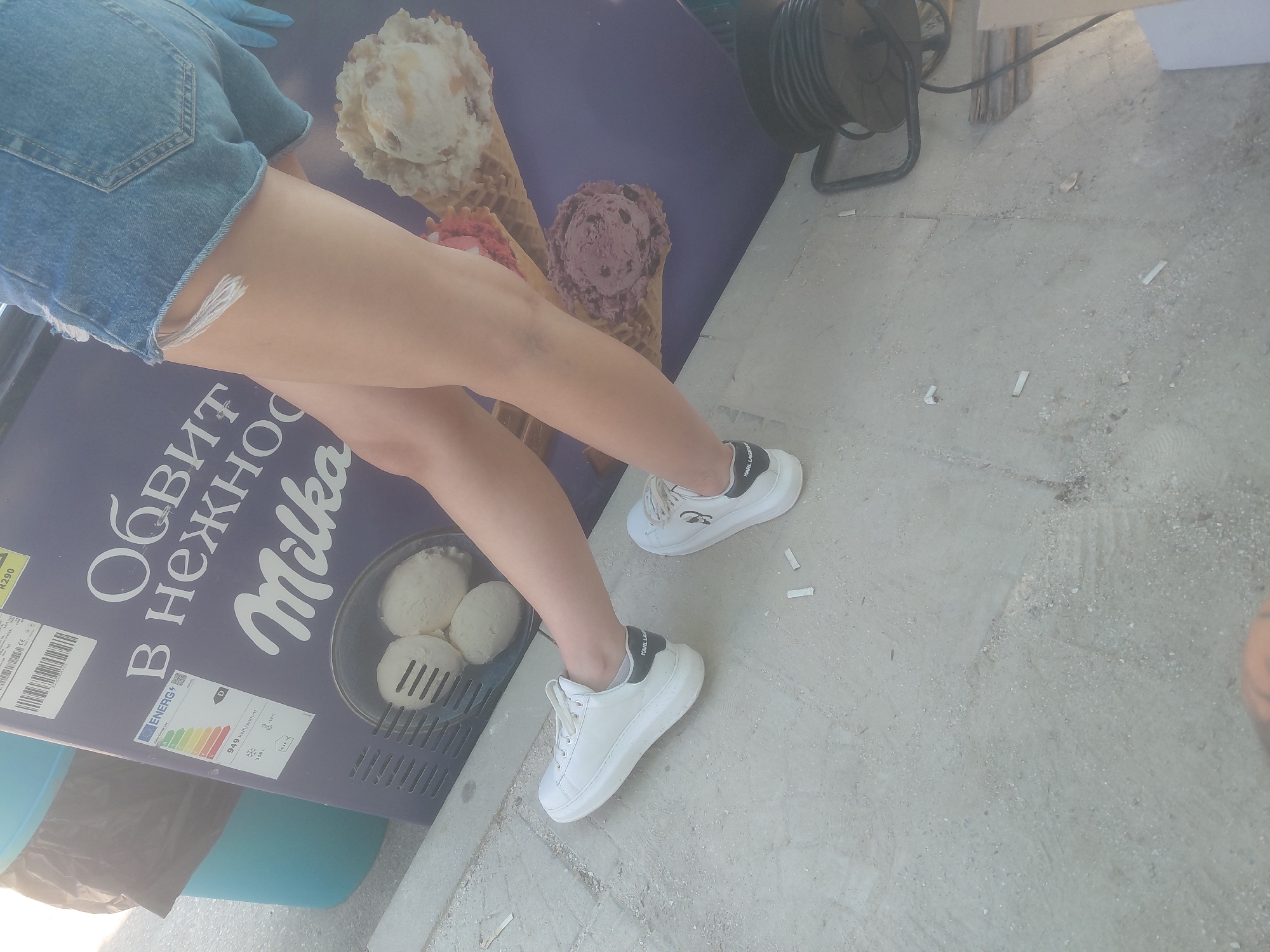 This image captures an outdoor scene in the city of Kostievo, Bulgaria, featuring the lower body of a person, a commercial ice cream display, and surrounding ground details.
The central subject is the lower torso and legs of an individual, dressed in light blue denim shorts with frayed edges and white platform sneakers. The left sneaker clearly displays "KARL LAGERFELD" branding on the black heel accent. The person is standing with their feet slightly apart, angled towards the left.
Adjacent to the person is a large, upright commercial ice cream freezer or display unit, predominantly purple in color. The unit features prominent white Cyrillic text, including the brand name "Milka" in its characteristic script font. Additional Cyrillic text, partially visible, reads "ОБИТ В НЕЖНО..." (Obit v Nezhno...), suggesting a phrase like "Coated in tender...". An energy efficiency label is affixed to the unit, displaying an energy class 'D' and indicating '949 KWh/annum'. Illustrated on the display are various ice cream items: three scoops of white ice cream in a metal container, and two large ice cream cones, one appearing to be vanilla or caramel flavored and the other a darker, possibly chocolate or berry, flavor.
The ground is a light gray concrete surface, appearing somewhat rough and uneven. Scattered across the concrete are several small, white cylindrical objects, identified as cigarette filter tips. In the upper left background, a light blue plastic bin containing a black trash bag is partially visible. To the far right, a black power cord reel is situated on the concrete, with electrical cords visible. The scene is brightly illuminated, suggesting daytime.