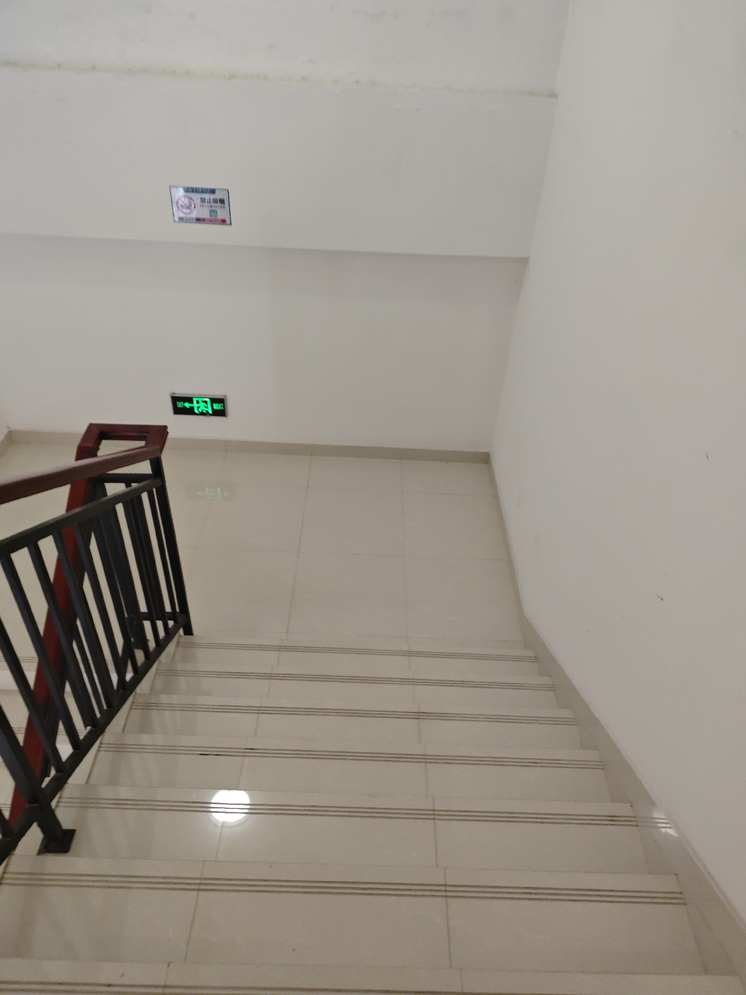 An indoor staircase with a dark metal railing and maroon handrail descends into the frame. The stairs are tiled with light-colored square tiles, each step having three dark parallel lines across its width. A green illuminated exit sign with Chinese characters is visible on the white wall above the staircase. Near the top of the wall, a small sign with a red circle and diagonal line over a cigarette symbol, indicating "No Smoking," is also present. The environment appears to be a building interior, possibly a public or commercial space, located in the Chongqing High-tech Zone, China.