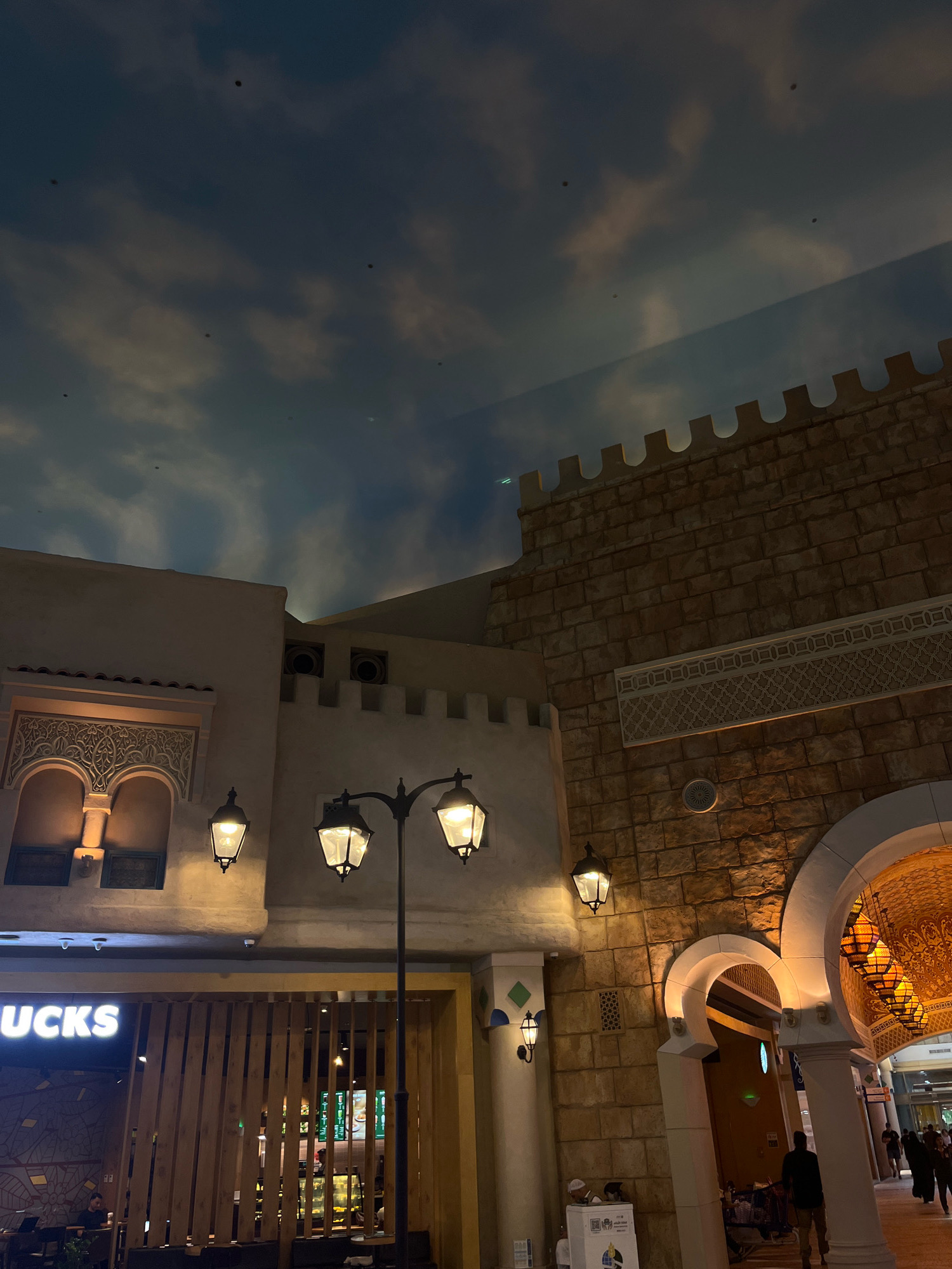 The image depicts an indoor scene designed to resemble a traditional Arabian marketplace or architectural style, likely within a shopping mall in Dubai, United Arab Emirates. The ceiling is painted to look like a cloudy sky. In the foreground, a Starbucks store is visible with its logo illuminated. To the right of the Starbucks, a section of a building constructed with stone blocks features decorative Islamic geometric patterns. An arched entrance leads further into the structure, revealing hanging lanterns. A street lamp with three lights stands in the middle ground. Several people are present, including a person sitting inside the Starbucks, and others walking in the background towards the arched entrance. The overall lighting suggests an evening or dimly lit indoor environment.
