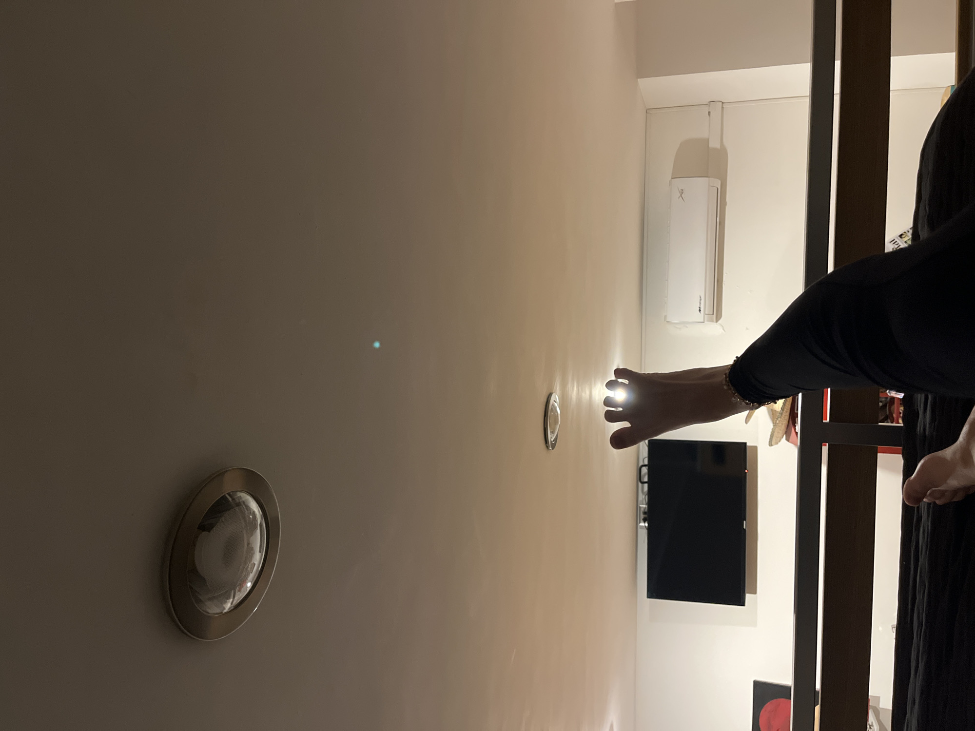 A low-angle indoor shot shows a wall with two ceiling lights, one at the bottom left and another near the center. A hand shines a light toward a television mounted on the wall. Next to the TV is a white air conditioning unit. A portion of a black garment and a shelf are also visible on the right. The image's location is unknown.