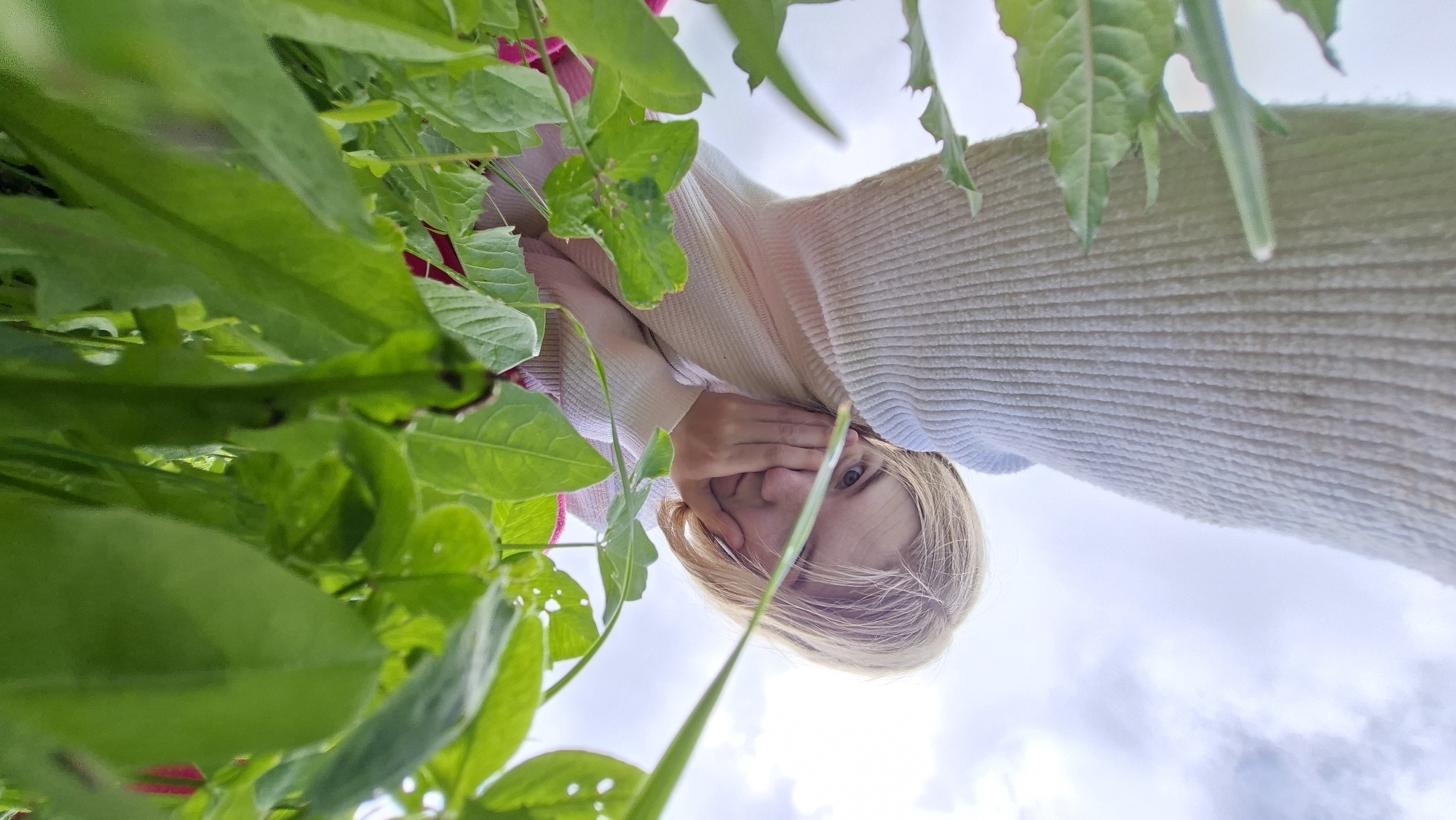 This image is captured from a low-angle perspective, looking upwards at a young woman with blonde hair. She is positioned above the camera, peering down, with her left hand gently covering part of her mouth and chin. One blue eye is clearly visible, conveying a gentle or possibly shy expression. She is wearing a light-colored, likely cream or off-white, ribbed long-sleeved sweater.
The foreground of the image is filled with lush green foliage, consisting of various leaves and plant stalks, some of which are softly blurred, creating a natural frame around the subject. This dense greenery suggests an outdoor setting, such as a garden or a field with overgrown plants.
Behind the woman, the background reveals a bright, overcast sky with visible clouds, indicating that the photo was taken during the daytime. The overall scene suggests an intimate outdoor portrait. There is no visible text in the image. The location context is Большой Букор, Russia.