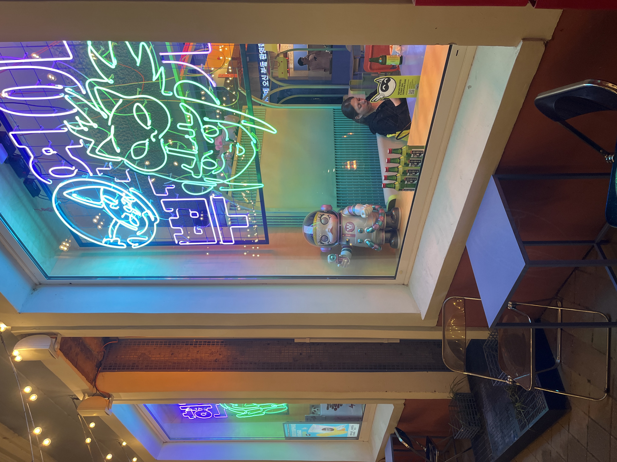 The image depicts the storefront of a business, likely a bar or restaurant, featuring bright neon signs in the windows. The signs showcase cartoon-like characters.
Inside the window, a person is visible in the reflection. A small robot figure is displayed on the windowsill. Bottles of a beverage are also displayed.
Outside the store, there is a table with chairs on the sidewalk. String lights are visible above the storefront. The overall setting suggests an urban environment, possibly Krasnodar, Russia. However, that cannot be verified solely from the image's content.