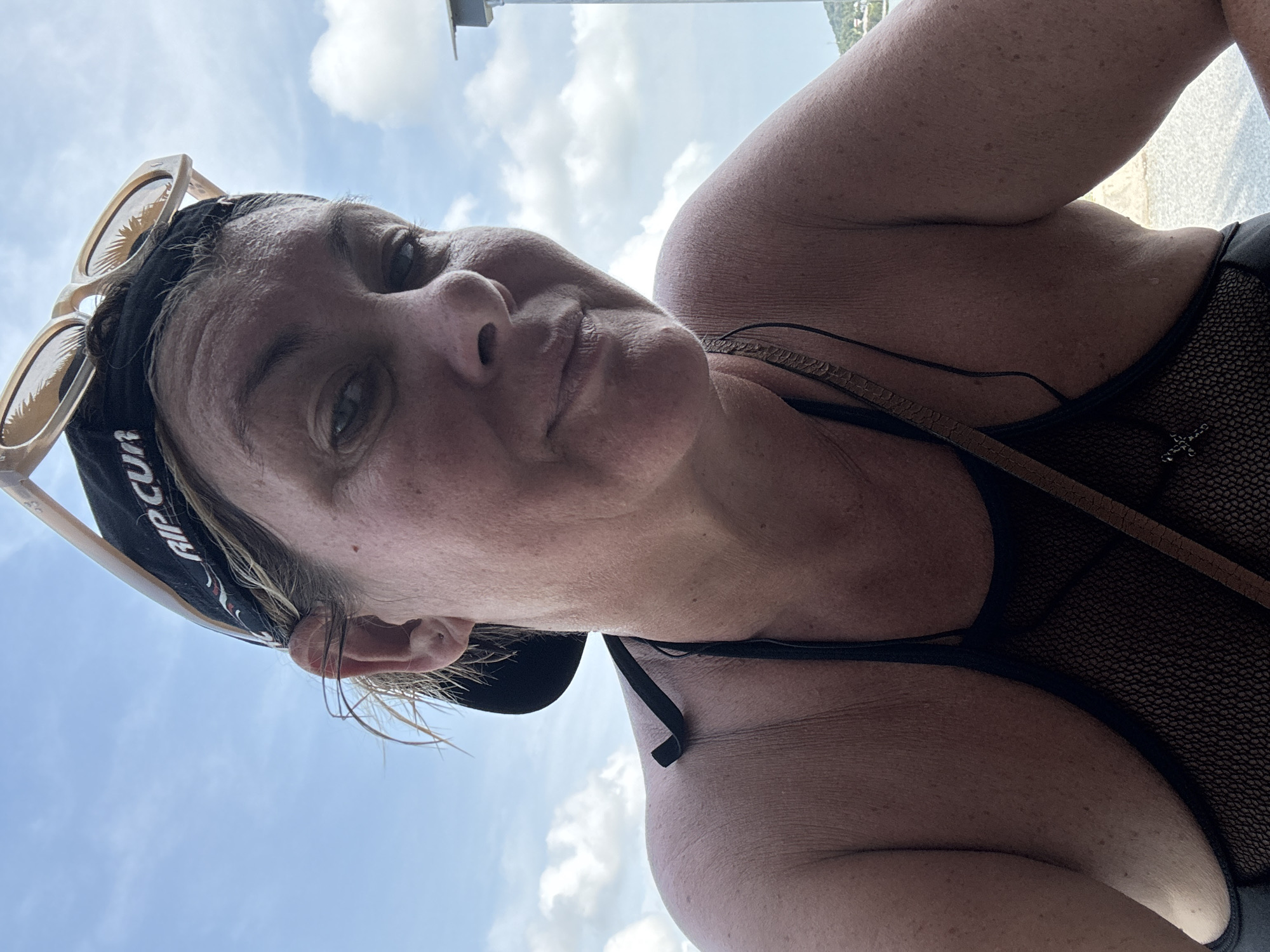 A woman is depicted from a low angle, looking down at the camera. She is wearing a black baseball cap with "RIP CURL" visible and sunglasses perched on her head. She has fair skin, and her facial features include visible freckles. The woman is wearing a black top with thin straps. The background features a clear blue sky with scattered white clouds. The location is indicated as Baan Chaweng, Thailand.
