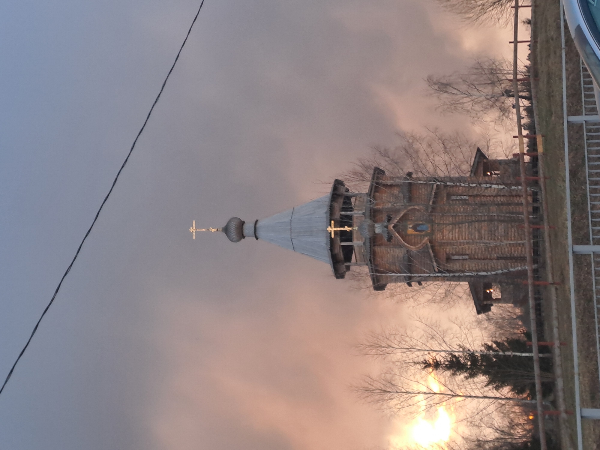 The image depicts a wooden church with a silver dome and cross, likely located in or near Vzglyadnevo, Russia. The church appears to be constructed of logs or timber, exhibiting traditional Russian architectural elements. The sky behind the church is a mix of gray and pale pink, suggesting either sunrise or sunset. Bare tree branches are visible around the church, indicating either late autumn or winter. A powerline crosses the upper left portion of the frame. A fence runs along the right side.