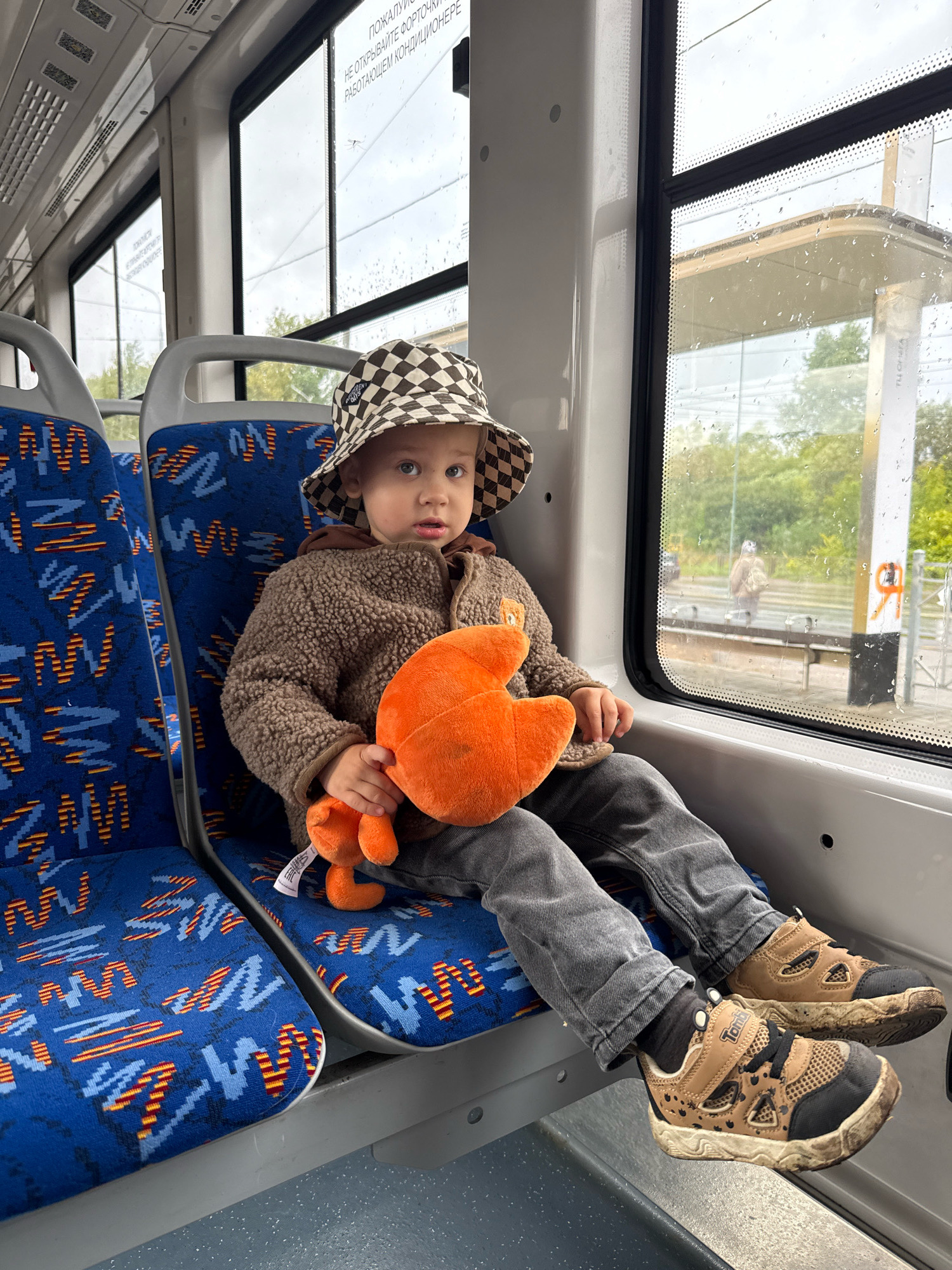 A young child wearing a checkered bucket hat, a brown fleece jacket, and gray jeans sits on a blue bus seat. The child is holding an orange plush toy and looking at the camera. The bus is moving, with a view of a bus stop and trees outside the window. The bus has patterned blue seats and a white interior. The scene appears to be in Yaroslavl, Russia, given the Cyrillic text visible on the window.
