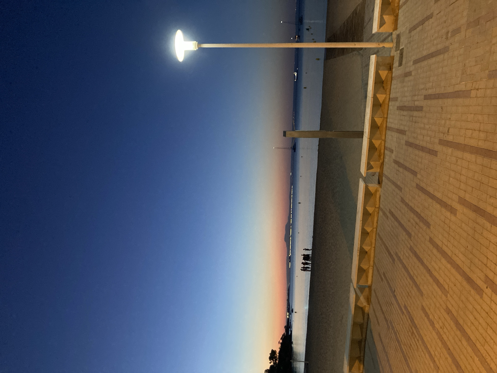The image depicts a beach scene at dusk in Palmanova, Spain. The sky transitions from a deep blue at the top to orange and pink hues near the horizon. A sandy beach occupies the middle ground, with the calm sea stretching out beyond. Several small boats are visible on the water. A group of people are standing together on the beach. In the foreground, a paved walkway is illuminated by a streetlamp on the right side of the frame. Several concrete benches with a yellow underside are also present along the edge of the walkway. The overall scene conveys a peaceful, end-of-day atmosphere.