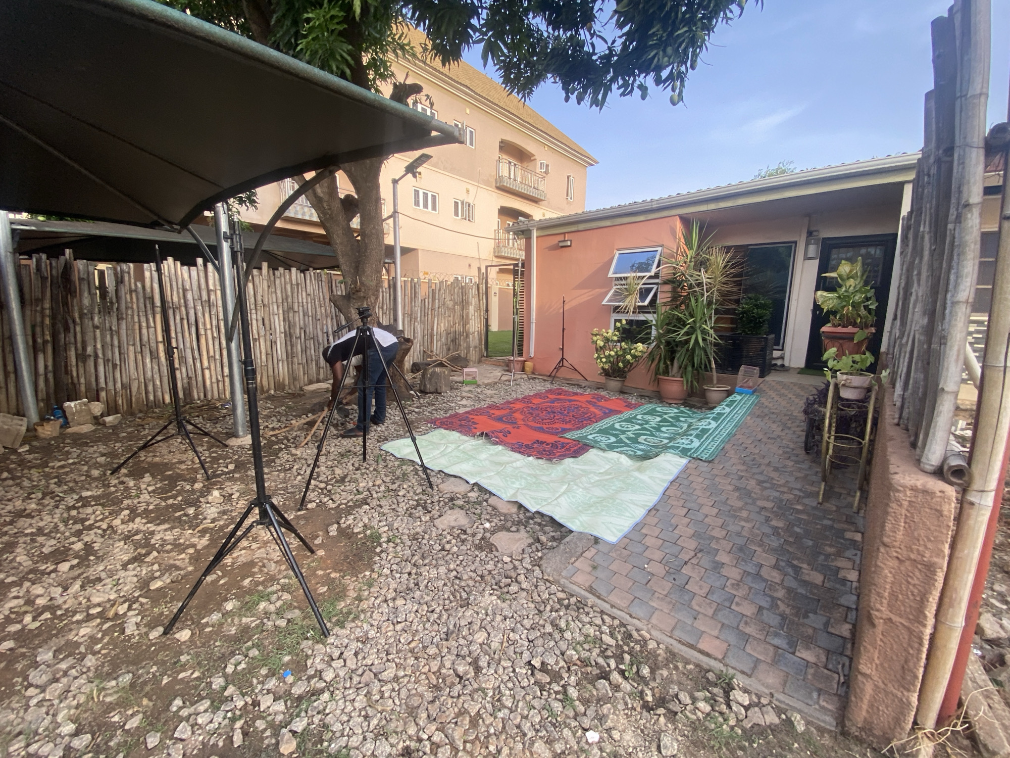The image shows a backyard setting in Barden Yana, Nigeria. The ground is a mix of gravel and brick pavers. A bamboo fence serves as a backdrop, partially covered by a large, dark-colored canopy held up by tripod stands. Several mats or rugs are spread out on the gravel area. A man is seen standing near a tripod, appearing to adjust equipment. A single-story building with orange-colored walls is visible. Windows with white frames are visible, along with potted plants near the building's entrance. A taller, multi-story building is visible in the background.