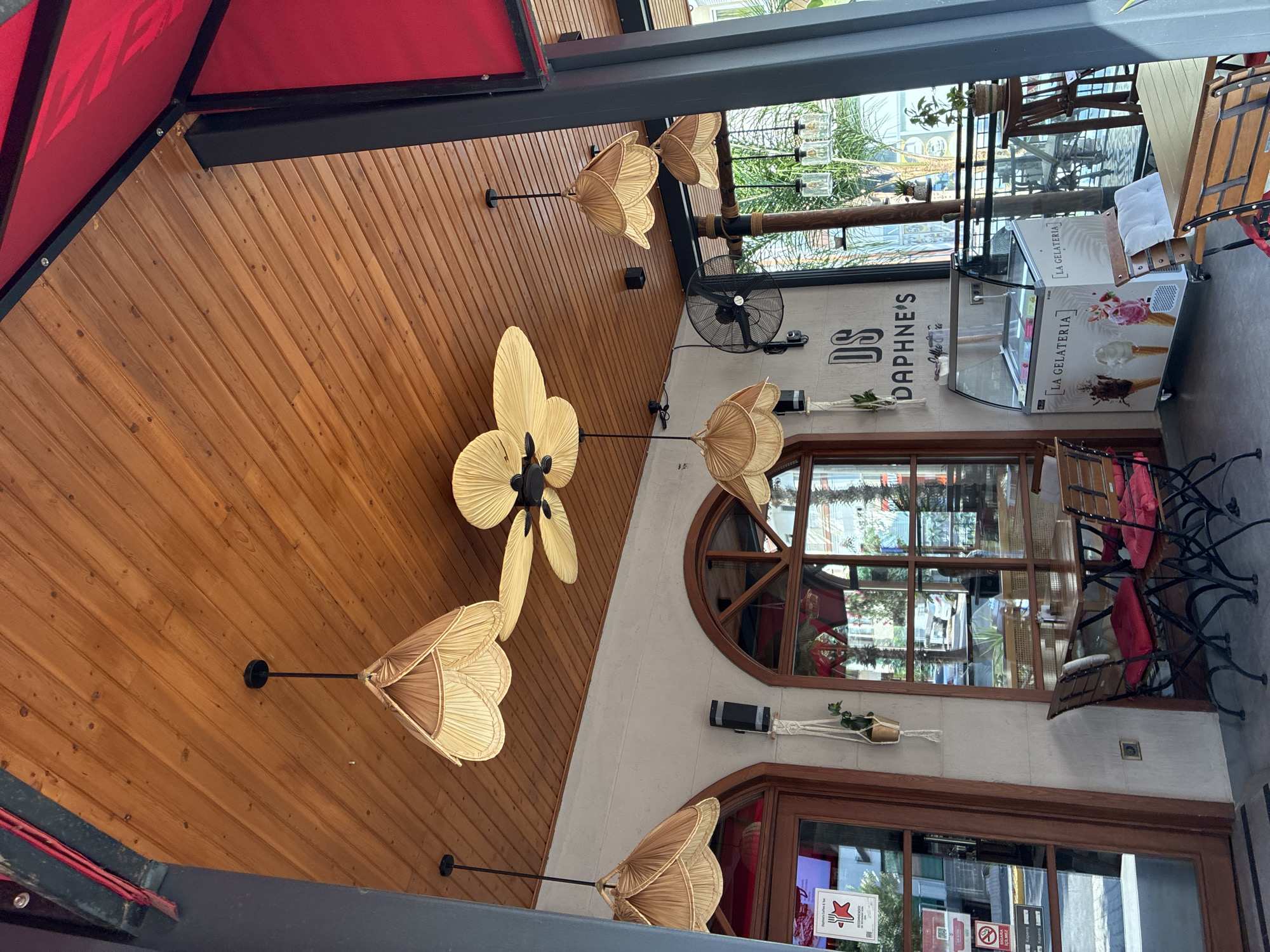 The image shows the outdoor seating area of a cafe or restaurant. The ceiling is made of wooden planks, and several decorative light fixtures with woven shades are hanging from it. A ceiling fan with petal-shaped blades is also visible. The walls are painted a light color, and there are arched windows with wooden frames. In the foreground, there are tables and chairs with red cushions. An ice cream display case with the words "LA GELATERIA" is positioned against the wall. The cafe appears to be located in Alanya, Turkey, as indicated by the "DS DAPHNE'S COFFEE & TEA" signage on the wall. The exterior environment visible through the windows suggests a sunny day with palm trees and other vegetation. There are no people clearly visible in the image.