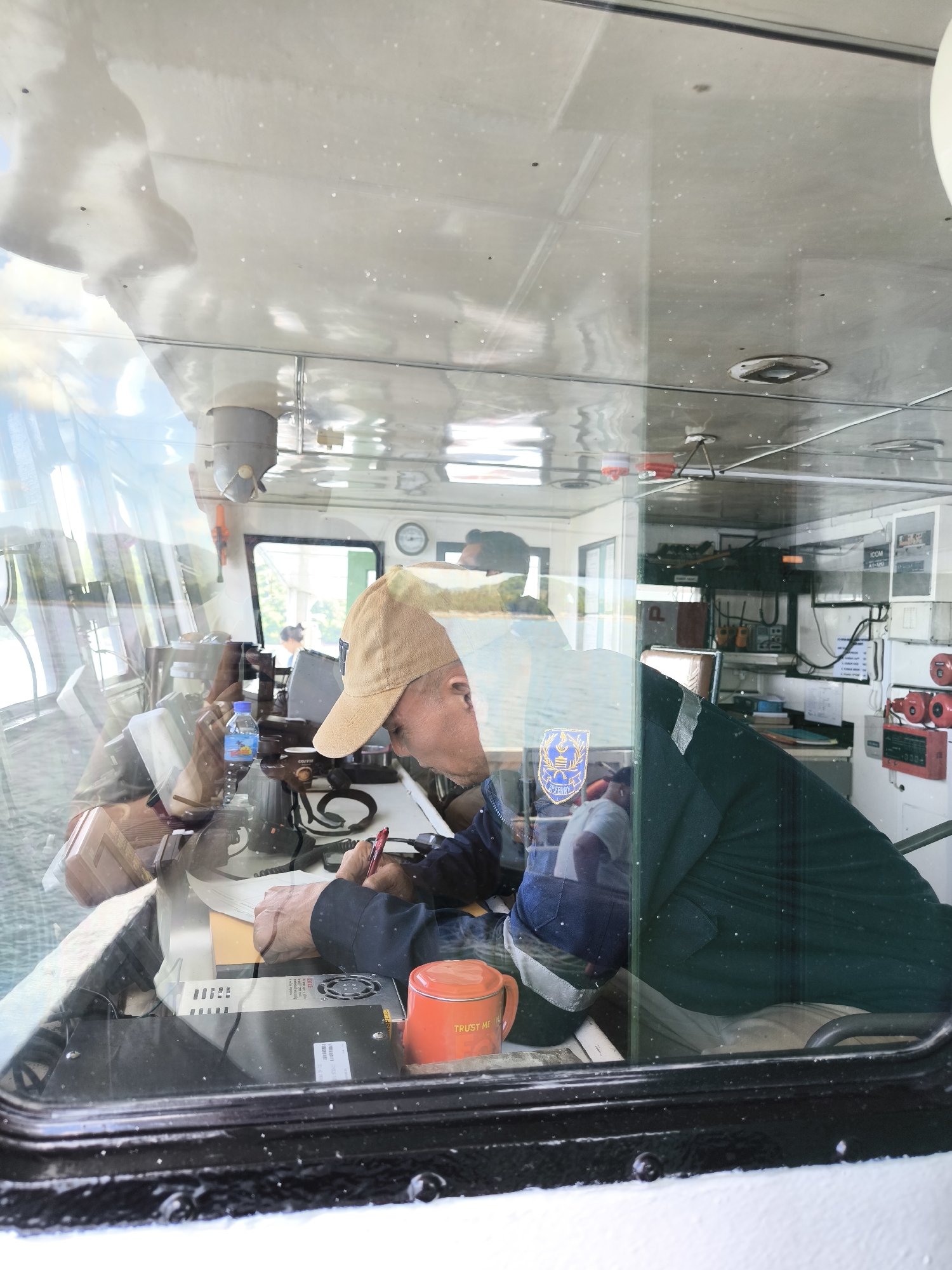The image provides a view into the pilot house of a boat or ferry, seen through a highly reflective glass window. A fair-skinned man, likely the captain or operator, is the central subject. He wears a tan baseball cap and a dark blue uniform shirt, actively writing on documents with a red pen at a desk. A blue water bottle, labeled "AIR MINUM" (Indonesian for "drinking water"), and an orange mug partially revealing the text "TRUST ME" are on the desk near his hands.
The glass creates strong reflections of the bright, cloudy sky and parts of the vessel's exterior, layering them over the interior scene. These reflections show overhead lights, ventilation, and other structures within the pilot house.
Behind the man, the control room is equipped with various instruments and communication devices, including an "ICOM AT-150" radio unit, clocks, and red fire alarm equipment mounted on the bulkhead. A blue and gold crest, possibly a company emblem for a "FERRY," is visible on what appears to be his uniform or a piece of equipment behind him. The scene depicts a busy control environment during daylight hours, consistent with operations in a location like Lembar Selatan, Indonesia. The man appears focused on his task.