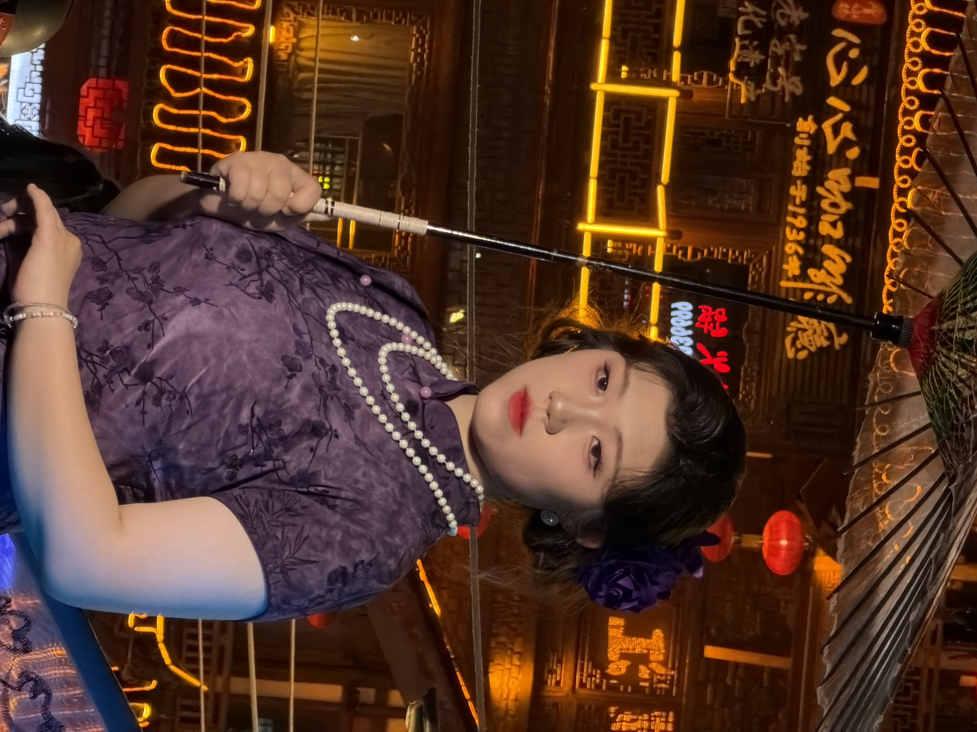 A young East Asian woman in traditional Chinese attire stands in what appears to be a historic district at night. She is holding a closed umbrella and wears a purple qipao with a floral pattern, a pearl necklace, a purple flower in her hair, and a bracelet. The background is illuminated by numerous neon signs with Chinese characters, suggesting a commercial area. The architectural style of the buildings in the background also points to a culturally significant location, possibly in Yuzhong District, China, known for its historical sites and vibrant nightlife. The overall scene evokes a sense of tradition and urban atmosphere.
