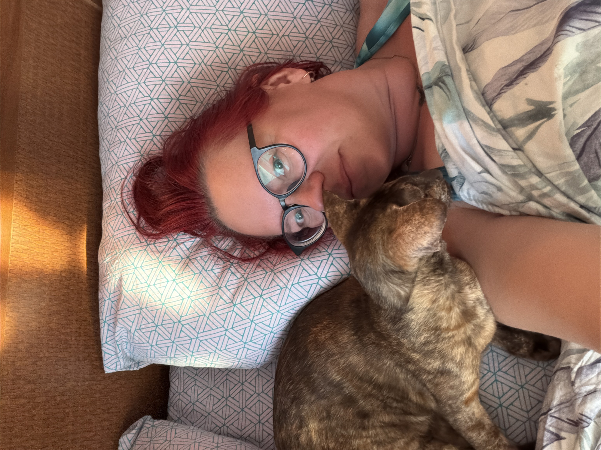 A woman with red hair and glasses lies on a patterned pillow in a bed, partially covered by a patterned blanket. A brown tabby cat rests on her arm. The location in the city of Chaikovsky, Russia is undetermined.