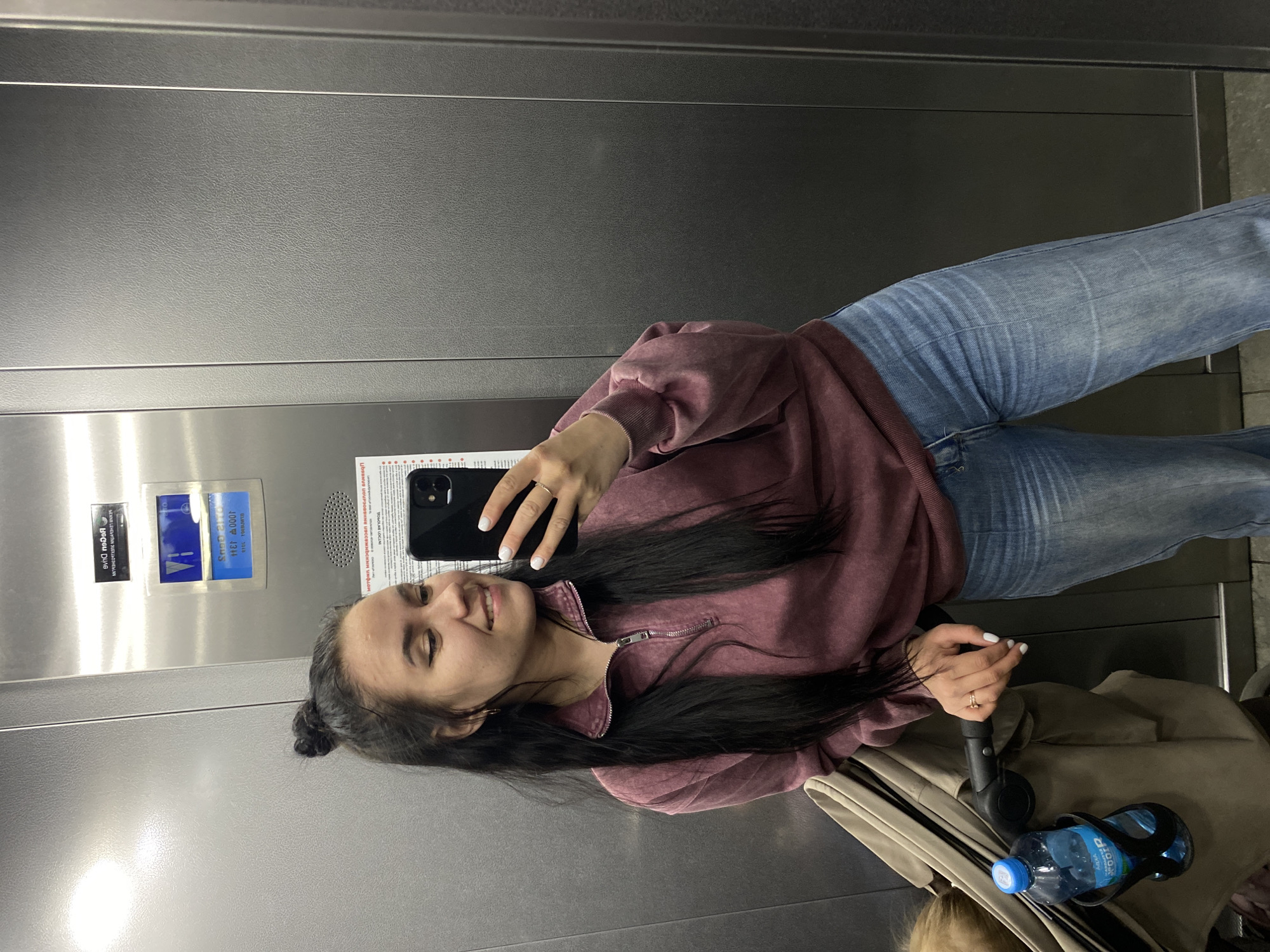 A woman is taking a selfie in what appears to be an elevator. She is wearing blue jeans and a mauve sweatshirt with a zipper closure. She holds a black smartphone in her right hand. Her long, dark hair is pulled back in a bun. Next to her is a stroller with a water bottle in a holder. In the background, there are informational signs written in Russian and containing the text: "BEGGU DUAG," "Snop 21," and "1000呎 134," along with other unreadable text. Based on the text on the informational signs, the location is likely in the city of Perm, Russia.