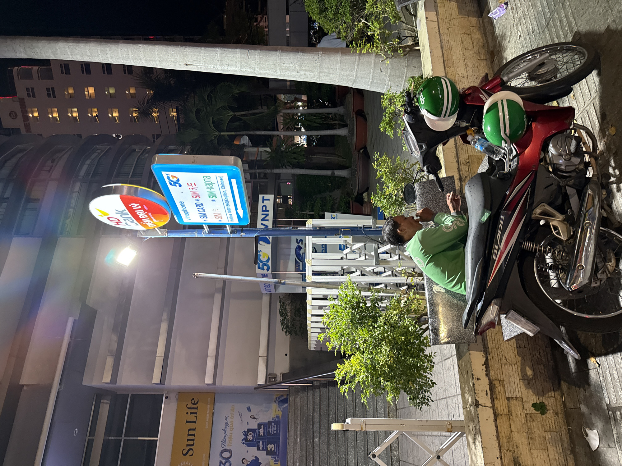 The image depicts an exterior scene at night in Nha Trang, Vietnam. In the foreground, a red and black motorcycle is parked. Two green and white helmets are attached to the handlebars, one on top of the other. A man wearing a green long-sleeved shirt is seated on a low wall to the left of the motorcycle, looking down and appears to be eating or smoking. Behind the motorcycle and the man, a row of lush green bushes is visible, bordering a textured stone wall.
Further back, a white fence-like structure stands, and behind it, a street lamp illuminates the area. A signpost with multiple signs is prominent. One sign is circular and red and white, displaying "HD Bank" and Vietnamese text. Below it, a rectangular blue sign features the Vinaphone logo and the text "5G" along with information about SIM cards in multiple languages, including English. Another sign beneath that reads "VNPT."
In the background, a tall building with numerous lit windows rises into the night sky. To the left of the main building, a large sign for "Sun Life" is visible. The overall environment suggests a commercial or public area within the city, possibly near a business or a service provider's location, given the presence of the bank and telecommunications signs. The lighting indicates it is after sunset.