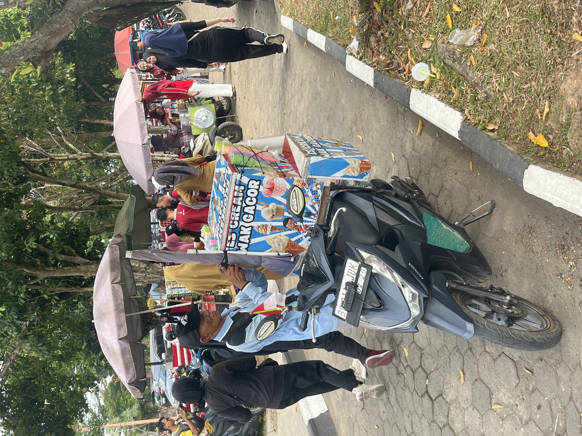 An outdoor scene in Sekayu, Indonesia, features an ice cream vendor with a cart attached to a dark-colored scooter. The vendor, an older man wearing a blue and black shirt, a black baseball cap, and a crossbody bag, is operating the cart. The cart is decorated with images of ice cream cones and text that reads "ES CREAM WAK GACOR." Several people are browsing or purchasing ice cream from other vendors under large umbrellas in the background. The ground is paved with hexagonal stones, and a grassy area with fallen leaves borders the paved area. The image appears to have been taken during daylight hours.