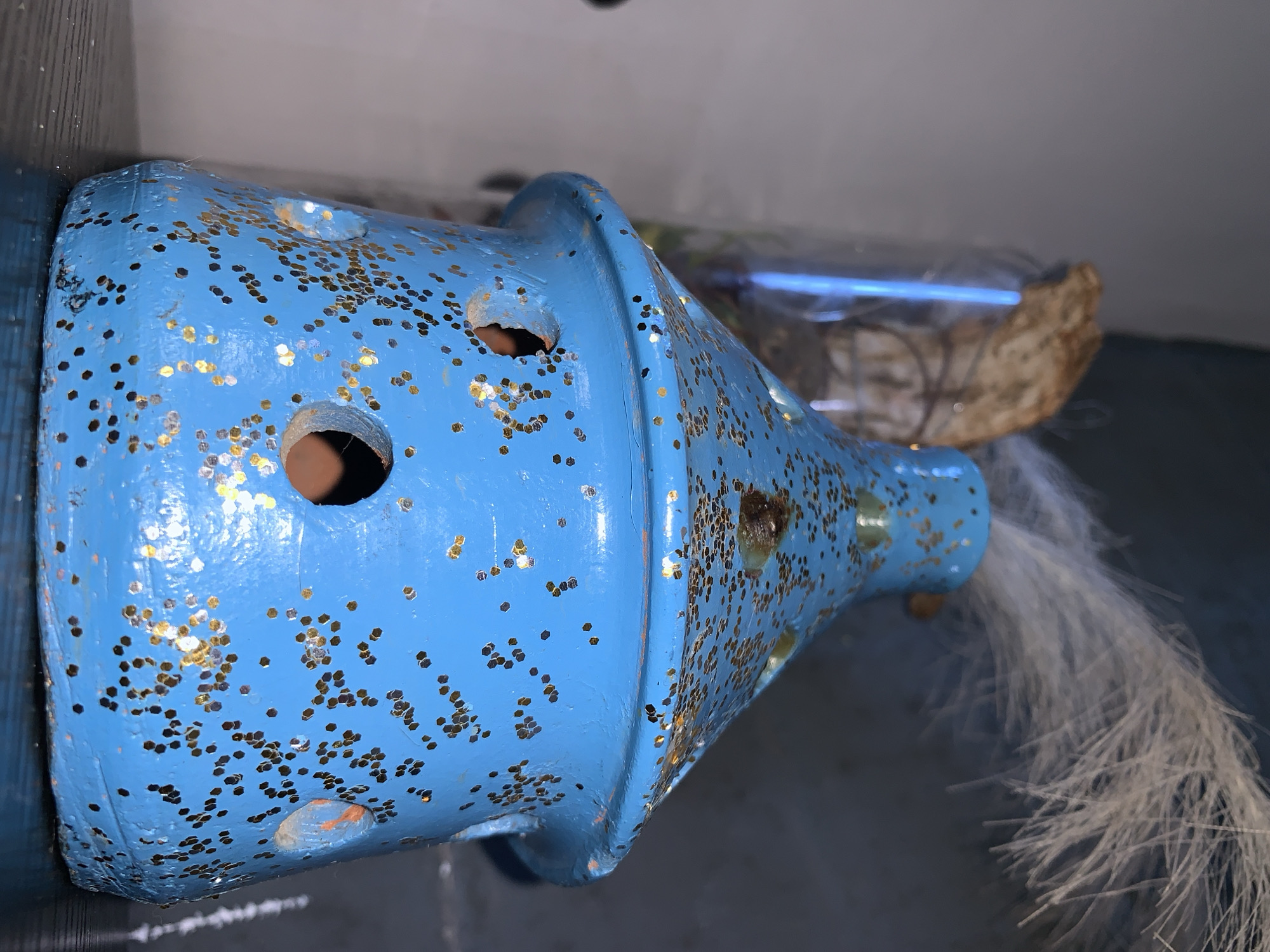 The image shows a close-up of a blue, glitter-covered, conical object, possibly a decorative birdhouse or incense burner. It has several circular openings and is adorned with gold glitter. A wispy white material is sticking out of the top. In the background, a glass container with a dried plant or branch is visible. The object is placed on a dark, textured surface, likely a shelf or table. There is no indication of the city of Jos, Nigeria, or any specific environmental details related to that location.