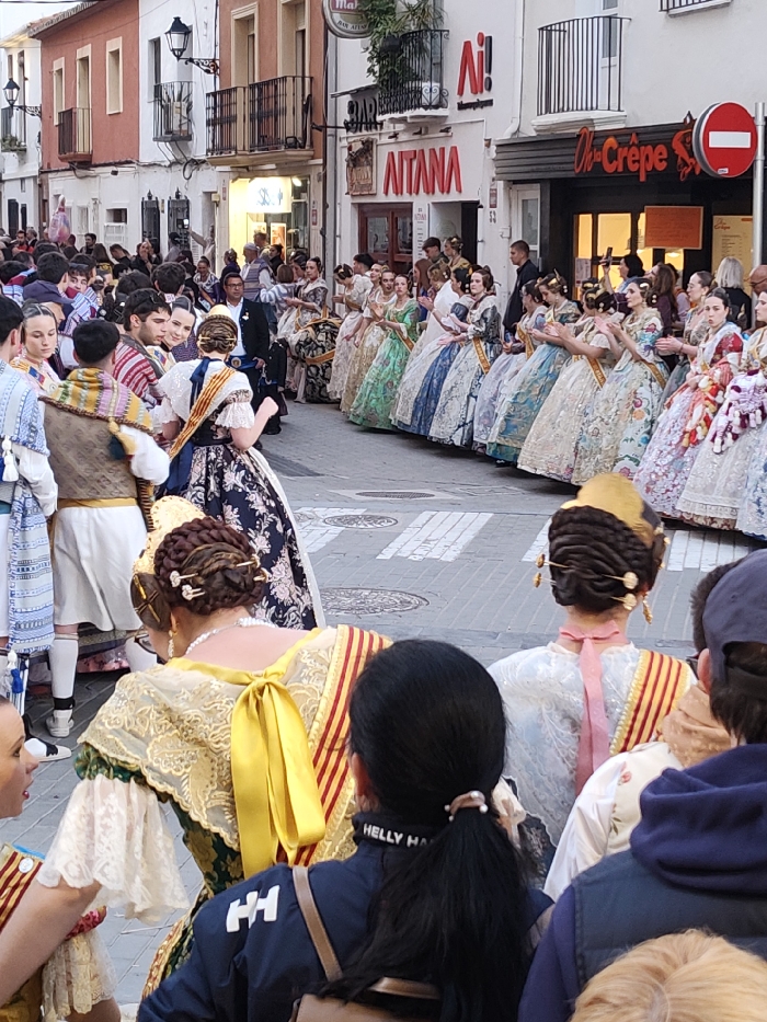 The image displays an outdoor street scene in Dénia, Spain, focusing on a large cultural gathering or procession. A prominent line of approximately two dozen individuals, primarily women, stands along the right side of a paved street. They are adorned in elaborate traditional Valencian "fallera" dresses, characterized by wide, patterned skirts, voluminous sleeves, and lace embellishments. Their hair is meticulously styled into intricate braided updos, frequently accessorized with decorative combs and pins. Many wear sashes across their chests, some featuring red and yellow stripes indicative of the Valencian flag. Several women in this line have their hands raised in front of them, possibly clapping or holding an unseen object.
In the mid-ground and left, additional participants in traditional attire, including men in vests and sashes, are intermingled with spectators dressed in casual clothing. The immediate foreground features the backs of several individuals, including a woman in a dark blue jacket with a "HELLY HANSEN" logo and another with dark hair in a ponytail.
The background consists of multi-story buildings lining both sides of the street, typical of Mediterranean architecture, featuring balconies with dark railings. Commercial establishments are visible on the ground floor, marked by signs such as "AITANA" in red lettering, "Ai!" in white, and "Oh-Crêpe Shop" with an orange sign. Adjacent to the "Oh-Crêpe Shop" sign, a red circular 'No Entry' traffic sign is mounted. Streetlamps extend from the building facades. White crosswalk markings and circular utility covers are visible on the grey street surface.
