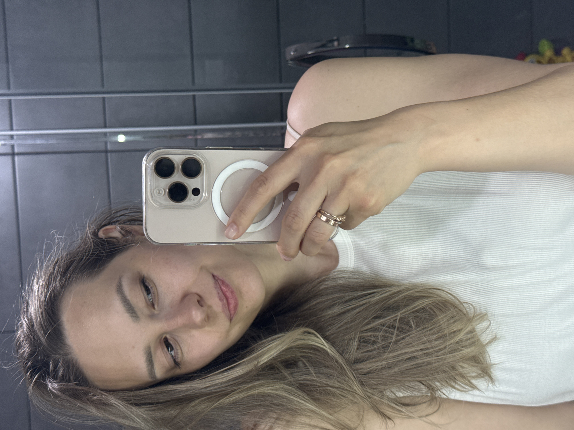 The image shows a woman taking a selfie in a bathroom. She is holding a smartphone with a clear case and a MagSafe ring attached to the back. The phone's camera is visible, featuring three lenses. She is wearing a white top and two gold rings on her finger. Her hair is blonde and styled loosely. The background includes grey tiled walls and a chrome towel rack. The location is likely a private residence bathroom, although no specifics regarding the city of Chaikovsky, Russia can be determined from the image.