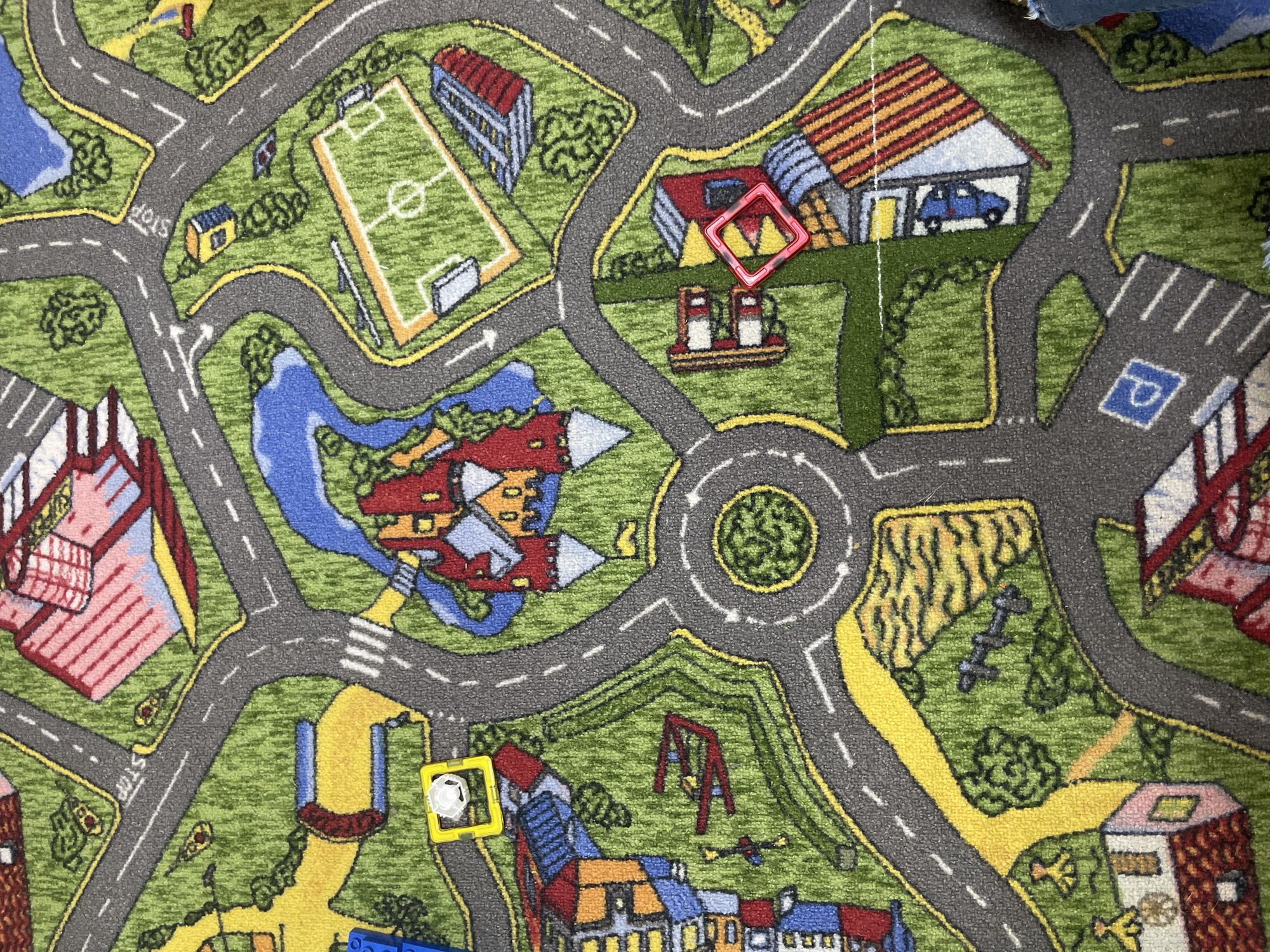 The image presents an overhead view of a children's playmat, intricately designed as a miniature town or city. A network of gray roads with white dashed lines and yellow borders dominates the scene, featuring intersections, turns, and a prominent roundabout in the center-right.
Various buildings and landmarks are depicted across the green, grassy landscape. These include multiple houses with red, orange, or striped roofs, one of which has a blue toy car parked inside its garage. A gas station with red pumps is visible, along with larger structures that resemble a school, apartment buildings, and a distinctive red and pink striped building. Recreational areas are also present, such as a soccer field with white markings and goals, a playground with red swings, and several blue ponds or rivers winding through the green areas.
Two toys are placed on the mat: a red magnetic tile with a geometric cutout sits on a road near the gas station, and a yellow square magnetic tile containing a small white cylindrical piece rests on another road in the lower-left quadrant. Blue Lego bricks are partially visible at the bottom edge of the frame.
Visible text on the mat includes "STOP" written in white letters at an intersection and a 'P' symbol indicating a parking area on a road. The image captures a static scene, suggesting a setup for imaginative play rather than an active event. The location context provided for this media file is Izhevsk, Russia.
