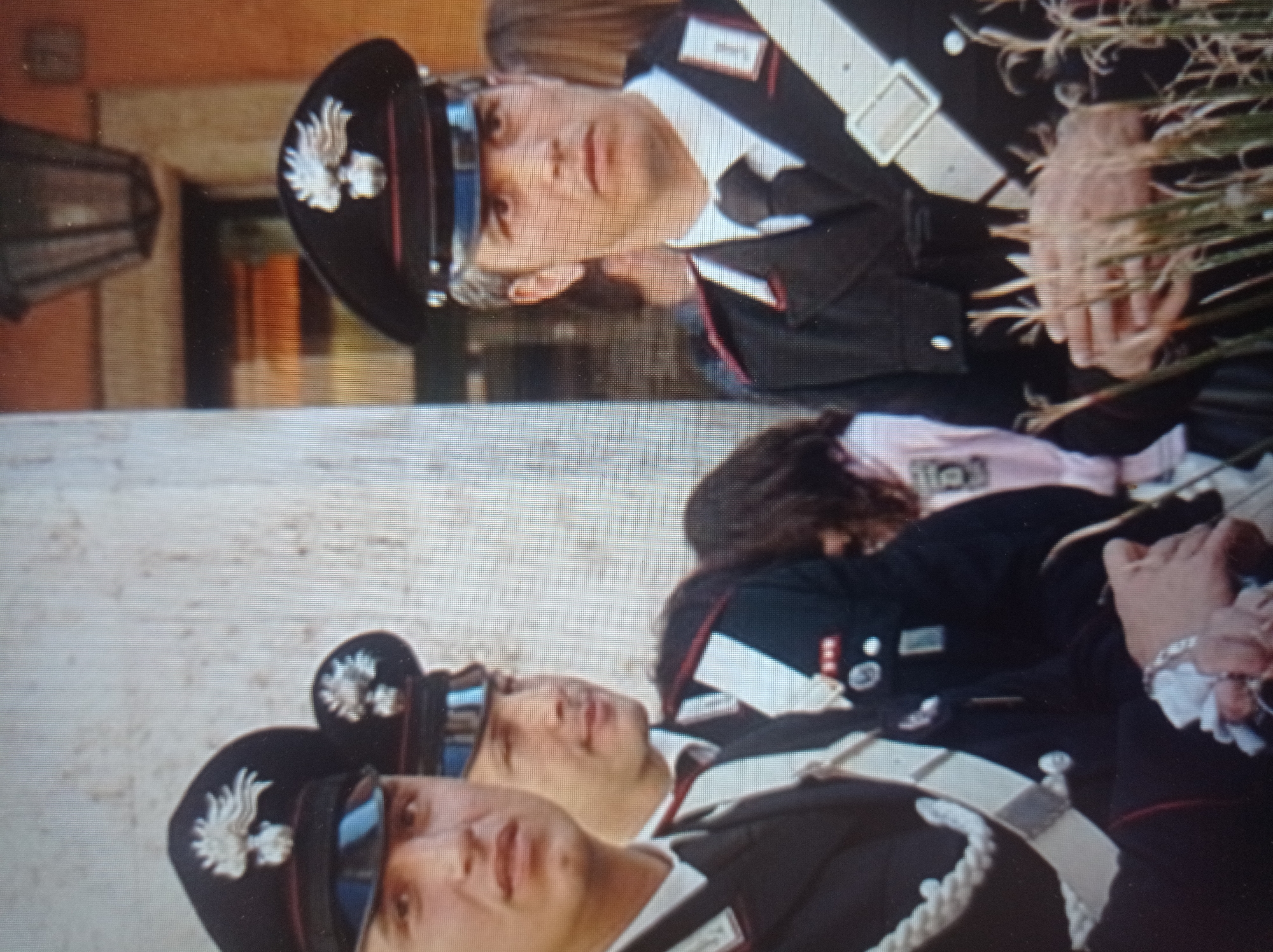 The image shows a group of men in uniform standing outdoors. The men are wearing black hats with insignia, dark jackets with white sashes across their chests, and ties. The uniforms suggest they are law enforcement or military personnel. One man in the foreground is looking directly at the camera with a serious expression. Another man to his left is partially visible, also in uniform. In the background, other people are blurred, and there is a building with architectural details and a lamp. The setting appears to be urban. The image does not provide any information about the city of Mukachevo, Ukraine.