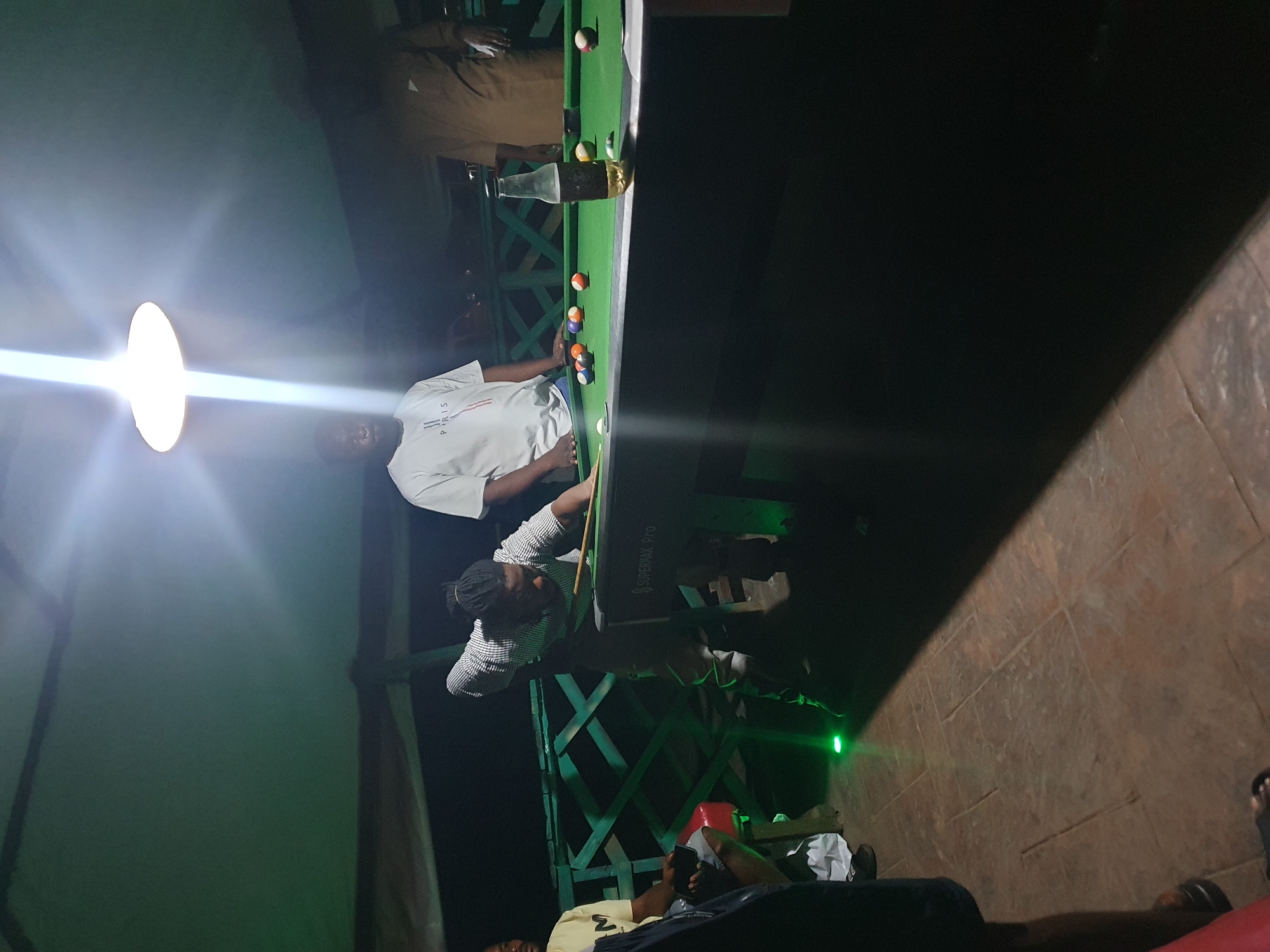 In Lokoja, Nigeria, a group of individuals gathers around a pool table under a covered outdoor area. The scene is dimly lit by an overhead light fixture, casting strong shadows. Three men are clearly visible: one in a striped shirt is actively playing pool, cue in hand, leaning over the table. Another man, wearing a white t-shirt with "PARIS" printed on it, stands near the table, watching. A third man, wearing a brown shirt, stands further back on the right, also observing the game. The pool table, labeled "SUPERMAX Pro," is green and has several colored balls scattered across its surface. A bottle of beer is standing upright on the table. The floor is paved with large, square tiles. A green railing is visible in the background, suggesting the area is elevated or part of a structure. The overall atmosphere appears casual and social.