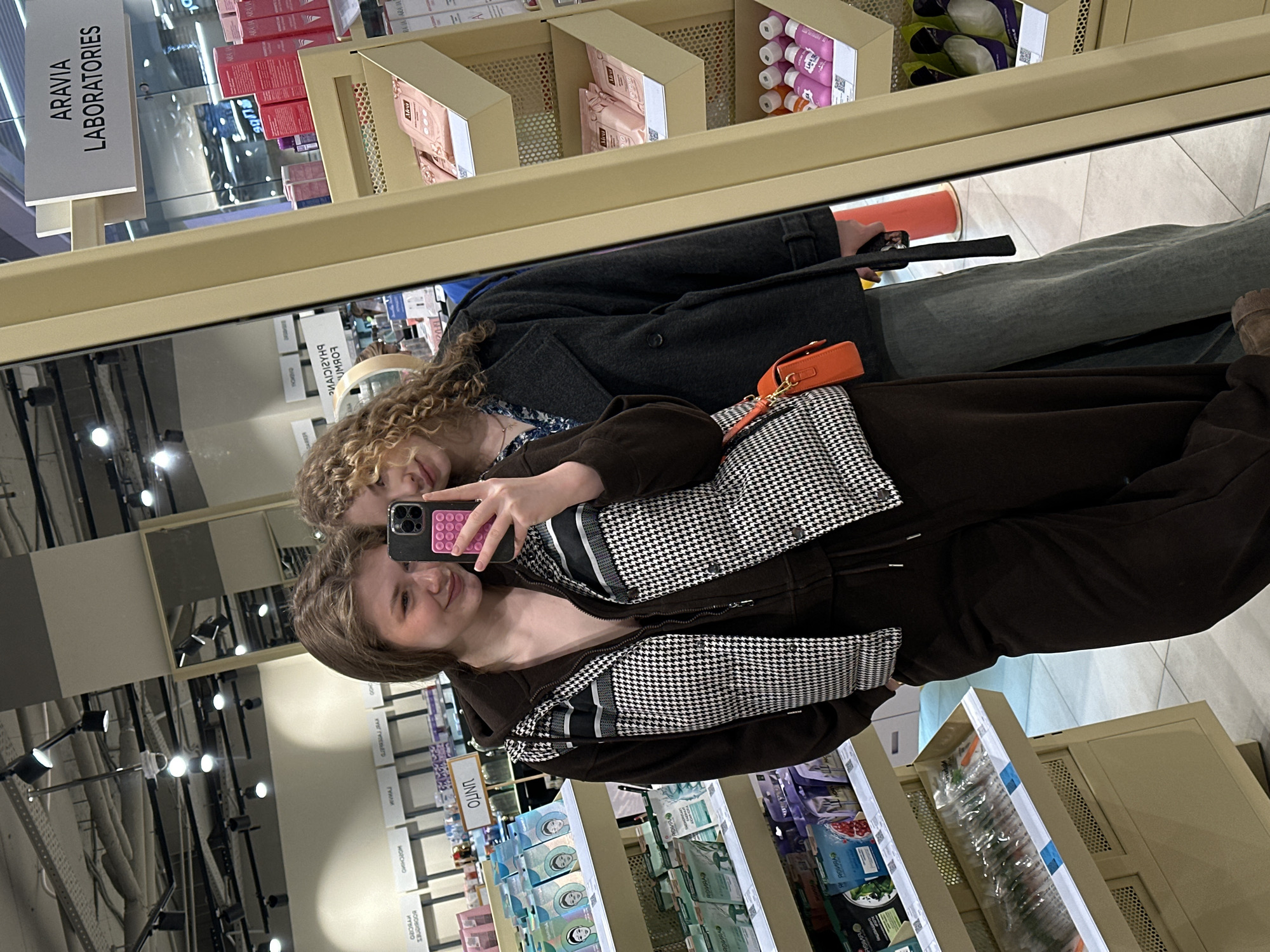A mirror selfie captures two young women inside a brightly lit retail store, likely a cosmetics or beauty supply outlet, located in Kazan, Russia. The woman on the left, with medium-length brown hair, smiles directly at the camera while holding a black smartphone, featuring multiple camera lenses and a pink pop-it grip, in her right hand. She is dressed in a dark brown hooded zip-up jacket worn open over a dark top, with a black and white houndstooth pattern sleeveless vest layered over the jacket. An orange small shoulder bag with a gold clasp is visible hanging from her right side. The second woman, with curly light brown hair, leans her head gently on the first woman's left shoulder, looking towards the camera with a subtle expression. She wears a dark gray double-breasted peacoat over a dark blue shirt with a light-colored floral pattern and light-colored wide-leg trousers. Her left hand is visible, holding a black strap or handle.
The background, reflected in the mirror, consists of multi-tiered retail shelving units stocked with various beauty and skincare products. To the left, shelves display numerous packaged face masks in green, purple, and white sachets. A backward-reflected sign above these products reads "ОЛИМП" (Olimp). To the right, adjacent to the mirror's light-colored frame, shelves hold pink boxed products and bottles with pink and purple liquids. A prominent sign above these products identifies "ARAVIA LABORATORIES." The store features bright overhead track lighting, exposed ceiling infrastructure, and a light-colored tiled floor. A red-orange cone-shaped object is partially visible on the floor behind the women. The setting is consistent with a beauty retailer, potentially within a shopping center such as TRC Olimp in Kazan.
