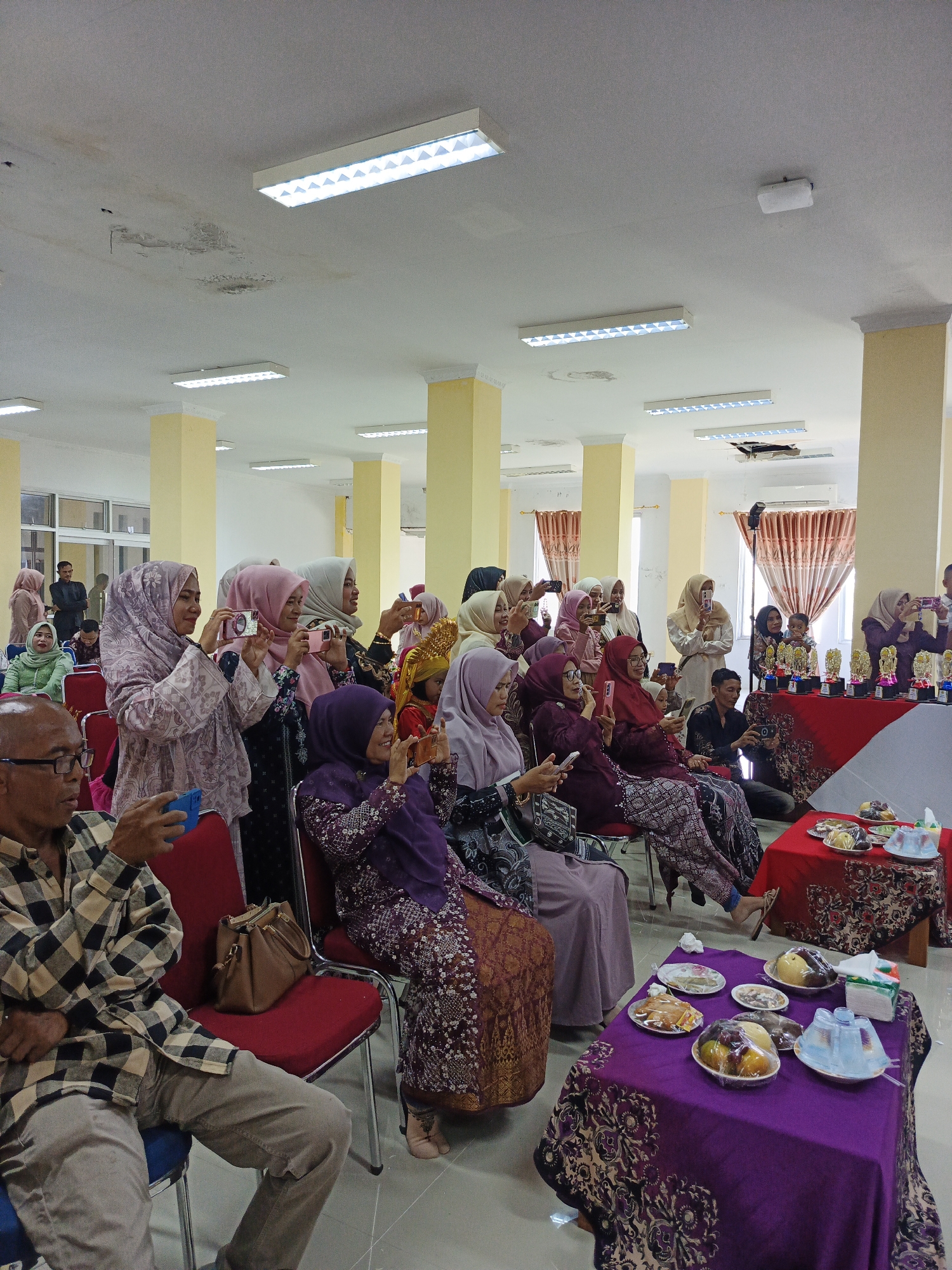 The image depicts an indoor gathering of approximately twenty people, predominantly women wearing hijabs and traditional attire, seated in rows of red chairs. Several individuals are holding up smartphones, appearing to take photographs or record videos of an event occurring out of frame. In the foreground, an elderly man with glasses is seated, holding a smartphone. To the right, a table is covered with a purple cloth adorned with floral patterns, displaying plates of fruit and snacks. Another table behind it, covered in a red cloth, holds several trophies. The environment appears to be a community hall or event space, characterized by visible ceiling lights and yellow support pillars. The overall scene suggests a formal or celebratory occasion. The lighting suggests it is daytime. Based on context provided by the image, the location is the Lhokseumawe City Hall in Lhokseumawe, Indonesia.