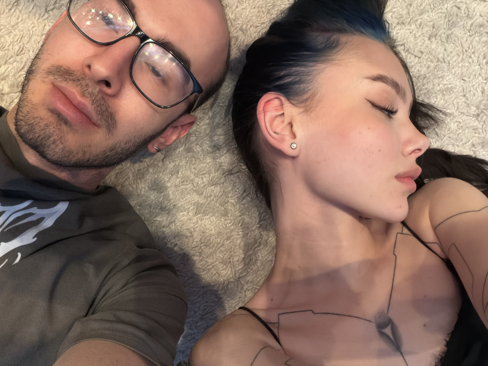 Two individuals, a man and a woman, are seen lying closely together on a light-colored, fluffy rug in an indoor setting in Khimki, Russia. Both appear to be resting or sleeping peacefully.
The man, positioned on the left, has short facial hair, including stubble and a light beard. He wears dark-rimmed glasses, which show reflections of lights. His left ear features a small hoop earring. He is dressed in a dark gray or greenish-gray hooded sweatshirt with a faint white design visible on his left shoulder.
The woman, on the right, lies with her eyes closed. Her dark hair shows streaks of blue or teal near the roots. She has winged eyeliner and visible blush. A small, round stud earring is visible in her right ear. Her chest and shoulder are adorned with intricate, fine line-art style designs, possibly tattoos or temporary body art, resembling a technical or schematic pattern. She is wearing a thin-strapped top or camisole.
The overall mood is calm and serene, suggesting an intimate moment of rest. No visible text is present in the image.