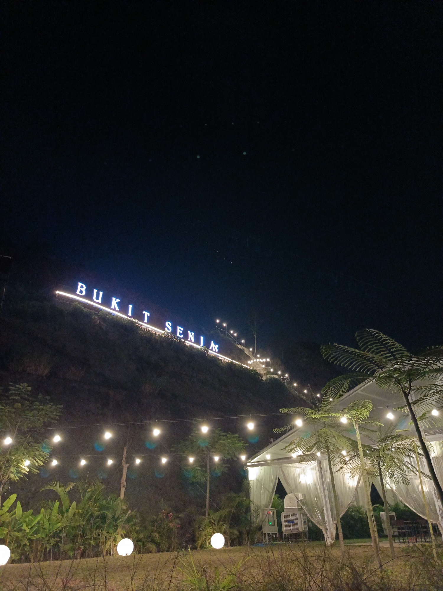 An outdoor nighttime scene in Cimahi, Indonesia, features a hill with large, illuminated blue letters spelling out "BUKIT SENJA" (Sunset Hill). The letters are mounted along the slope of the hill, which is covered in vegetation. String lights, also illuminated, are visible on the hill, ascending towards the sign. In the foreground, a white tent structure with translucent walls is set up, illuminated from within by warm, yellow lights. Several tall, thin palm-like trees stand in front of the tent. Additional round, white globe lights are placed on the ground near the tent. The sky is dark and clear, with only a few faint, scattered stars or distant lights visible. The area appears to be a park or an event space, decorated with lights for an evening gathering.
