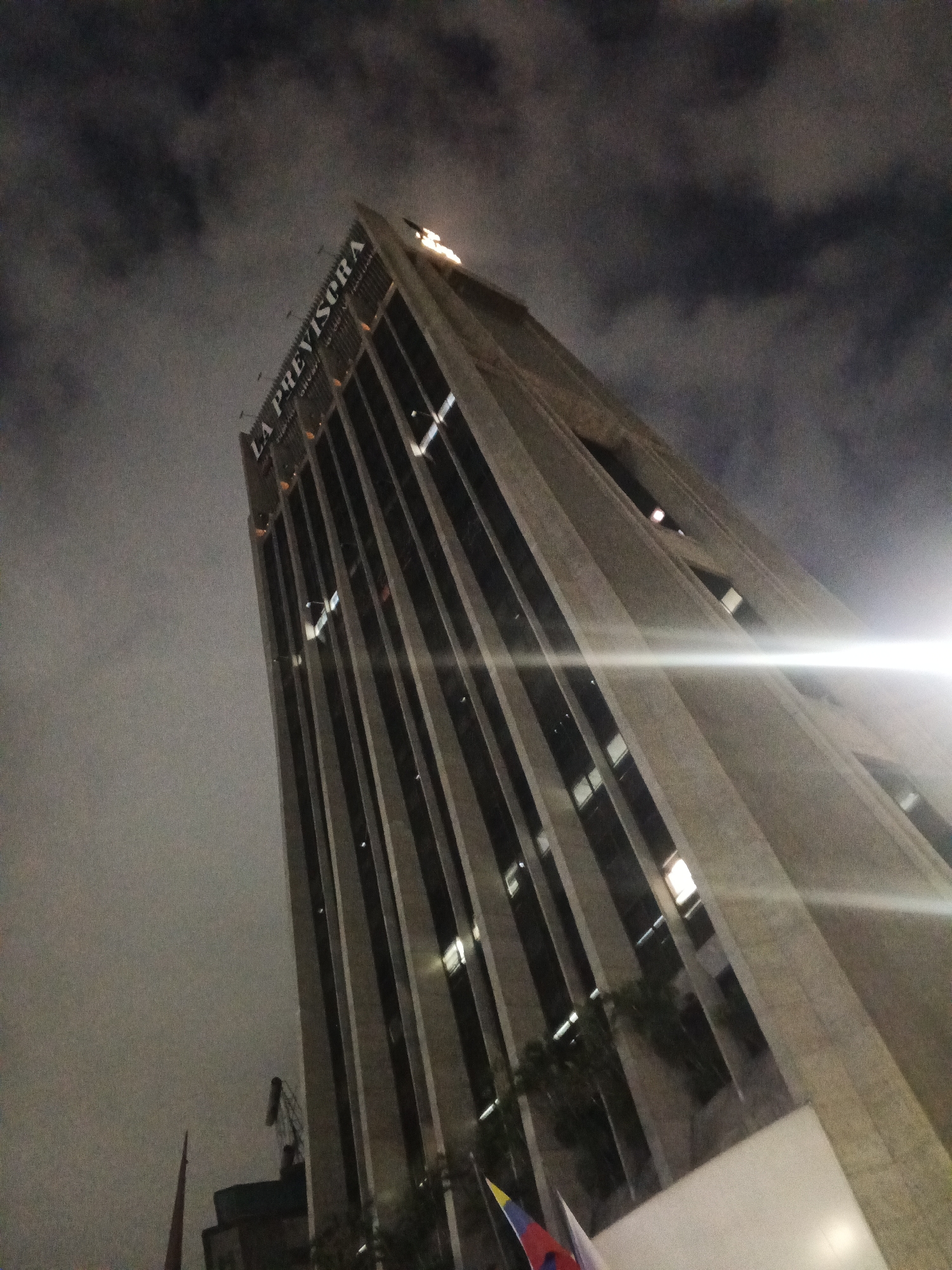 The image shows a low-angle, upward-looking view of the "La Previsora" building at night in Caracas, Venezuela. The sky is dark and cloudy, with some ambient light reflecting off the building's facade. The building itself is a tall, modern skyscraper with a distinctive angled design, featuring vertical sections of darker glass and lighter concrete or stone. Some windows on the building are illuminated, indicating activity within. A bright light source, possibly a streetlamp or a vehicle's headlights, creates a strong glare across the middle of the image, obscuring some of the building's details. In the lower left portion of the image, the top of a red structure, possibly a crane or a spire, is visible. There are also hints of flags and other buildings in the immediate foreground. The overall impression is of an urban nighttime scene with a focus on the architecture of the prominent La Previsora building.
