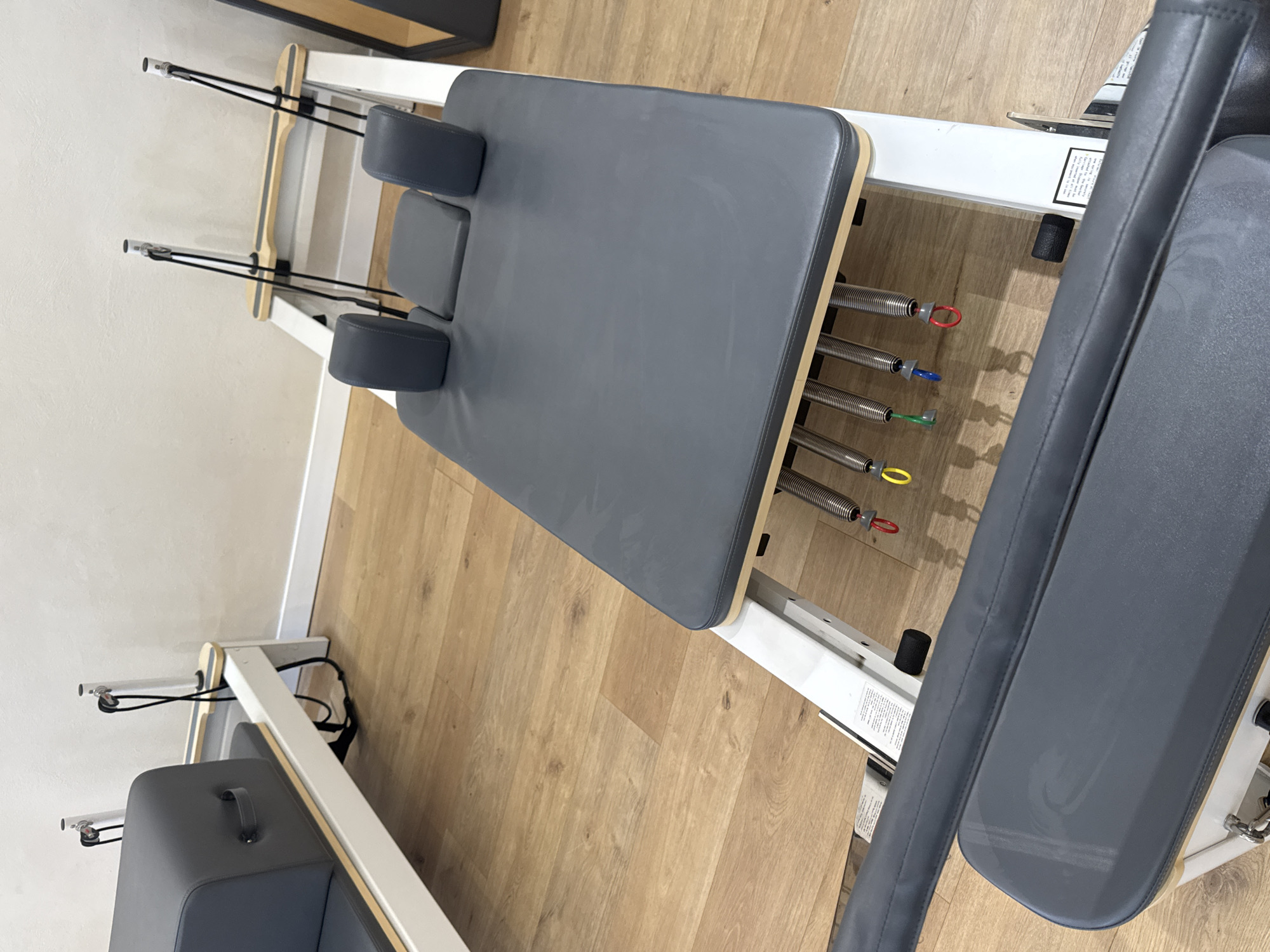 The image depicts a Pilates reformer machine, positioned on a light brown, wood-look floor. The reformer features a gray, rectangular carriage with padded headrests and shoulder blocks. Springs of varying colors are attached to the carriage on one end. The reformer frame is white and wood-colored. In the background, a portion of a white wall is visible, along with a small section of a black table or shelf. The image was captured in an indoor setting, likely a Pilates studio in Ramat Gan, Israel.