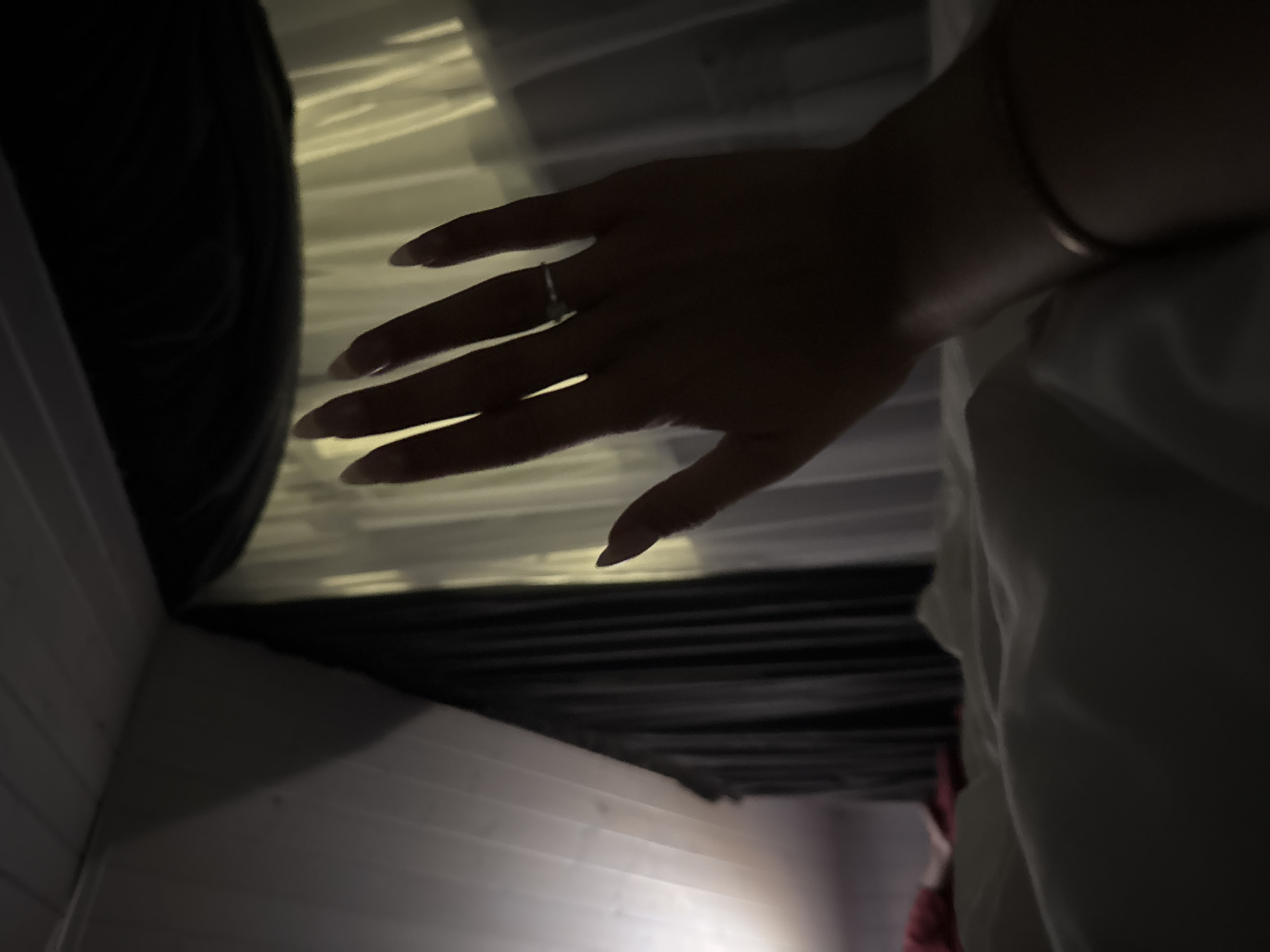 A low-light, high-angle shot shows a person's left hand, palm facing outward, against a backdrop of sheer curtains illuminated by an unseen light source. The hand is adorned with a ring on the ring finger and a bracelet on the wrist. The bed linens in the foreground are soft and rumpled, suggesting a relaxed or intimate setting. The scene is framed by dark fabric and a white wall with a textured ceiling, possibly in a bedroom. The location is identified as Doma 1096 Km, Russia.