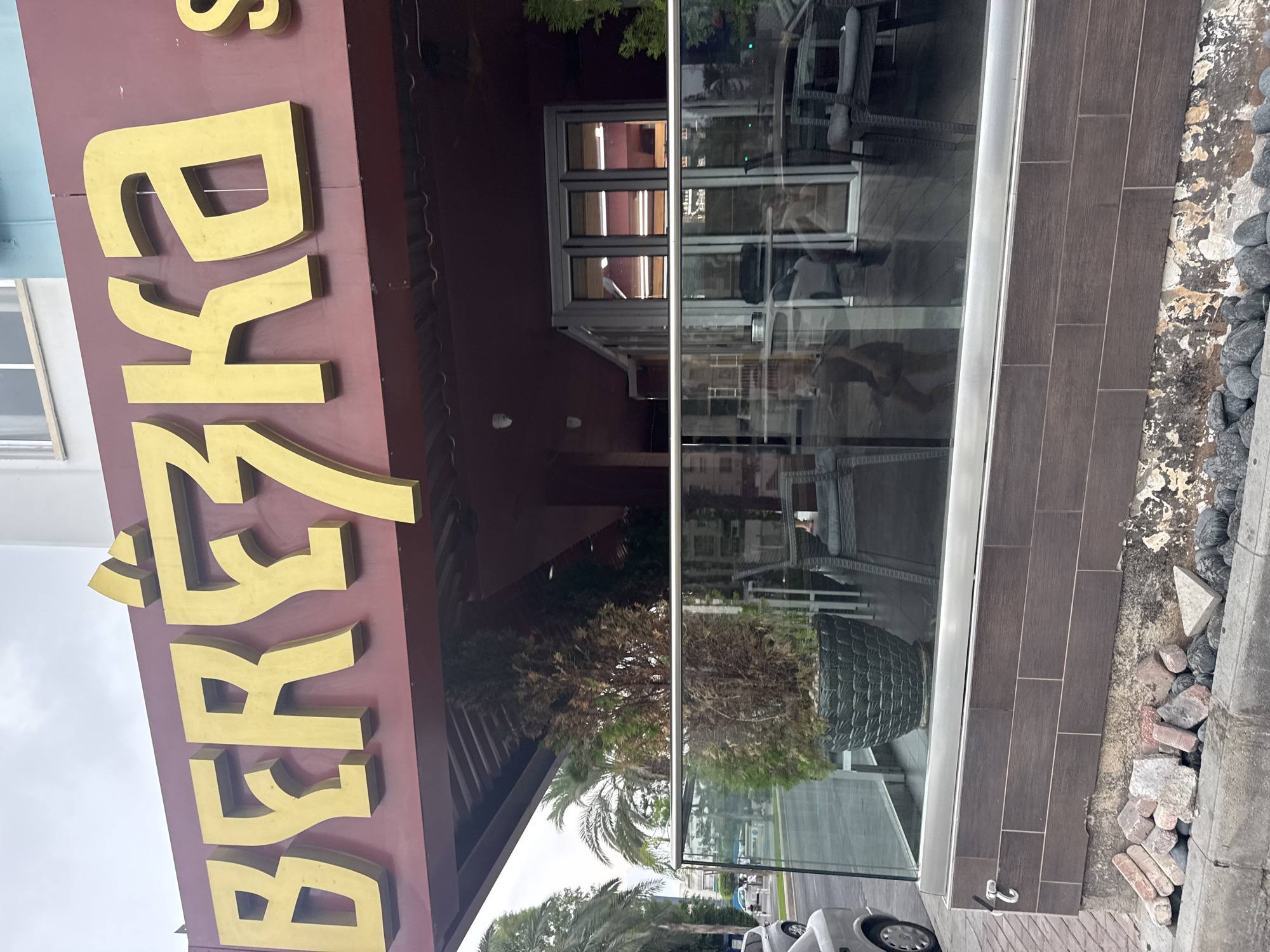 The image shows the exterior of a restaurant named "BEREZZKA" located in Germasogeia Municipality, Cyprus. The restaurant has large, golden, three-dimensional lettering spelling out its name on a maroon awning. In front of the restaurant, there is an outdoor seating area with glass railings, tables, and chairs. A tall potted plant with dried foliage is visible near the entrance. The street in front of the restaurant is paved, with a curb and some loose stones and debris. Cars are visible in the background, suggesting a public area. The sky is overcast.
