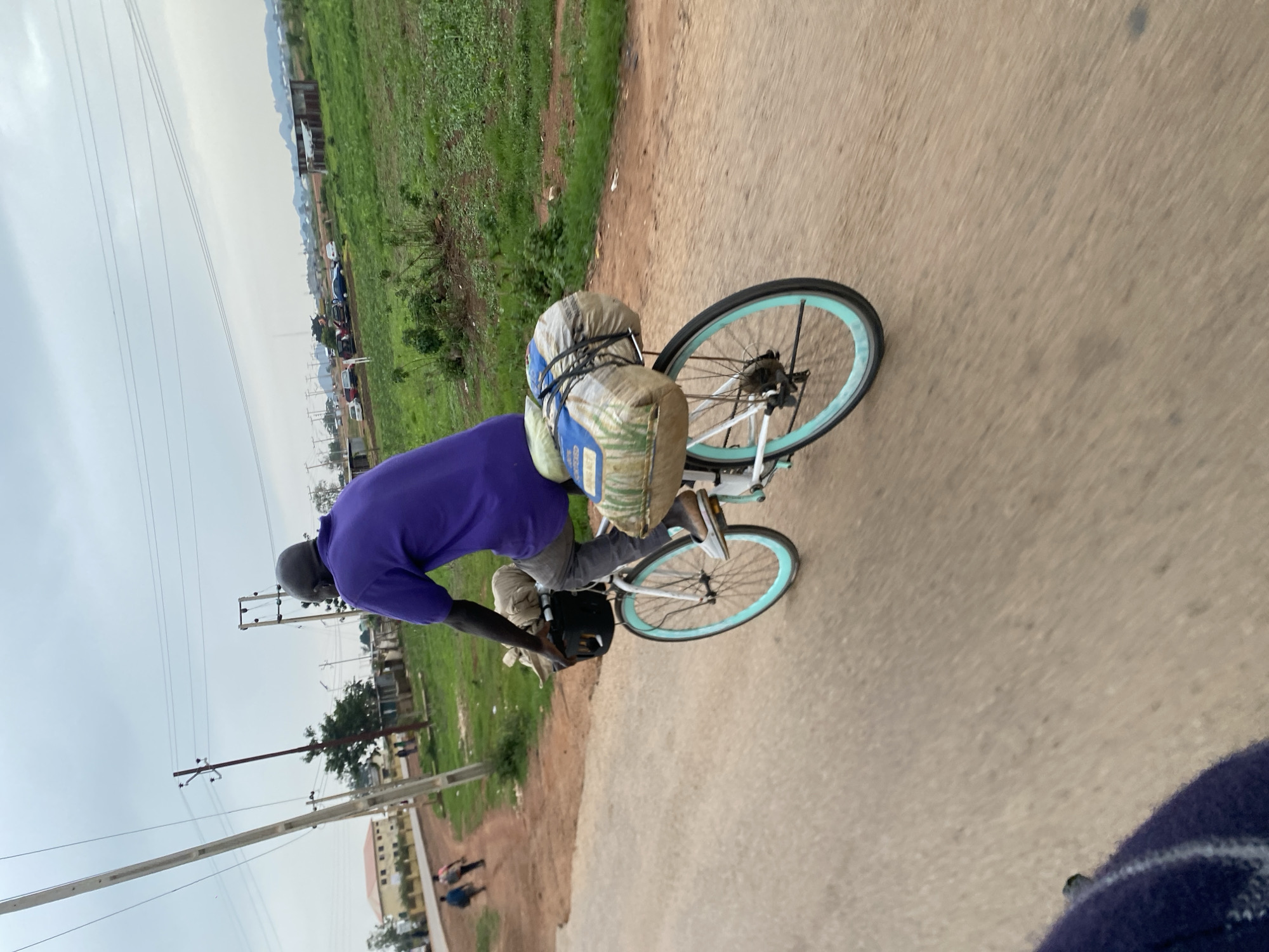 A person wearing a purple shirt and grey pants rides a bicycle on a dirt road in Jos, Nigeria. The bicycle is loaded with two large sacks. The background shows green fields, buildings, and utility poles. The sky is overcast.
