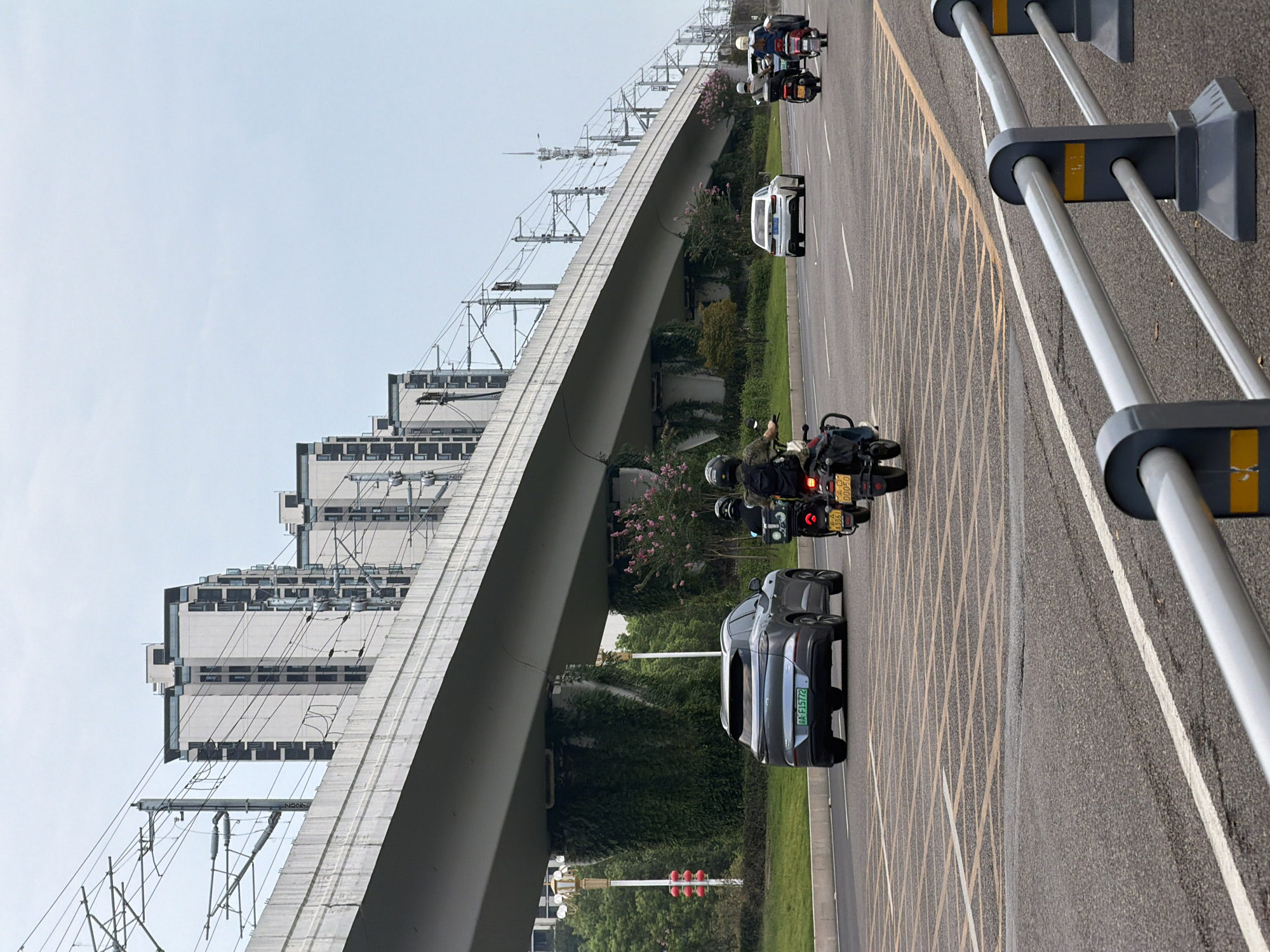 The image depicts a street scene in Jiangjin, China, characterized by a prominent elevated roadway or bridge structure overhead, lined with overhead power lines for what appears to be a rail system. Below the elevated structure, a multi-lane asphalt road is visible, with traffic comprising a gray SUV, two motorcycles with riders, and a white sedan. The SUV, bearing a license plate reading "A F15772," is in the left lane. The motorcycles are in the right lane, with the lead motorcycle carrying two individuals and the following motorcycle carrying one. The road is marked with yellow and white lane lines, and a pedestrian crossing with yellow box markings is present. Buildings, some appearing to be apartment complexes, are visible in the background behind the elevated structure. Greenery, including ivy covering the pillars of the elevated structure and trees along the roadside, adds to the urban landscape. The sky is overcast.