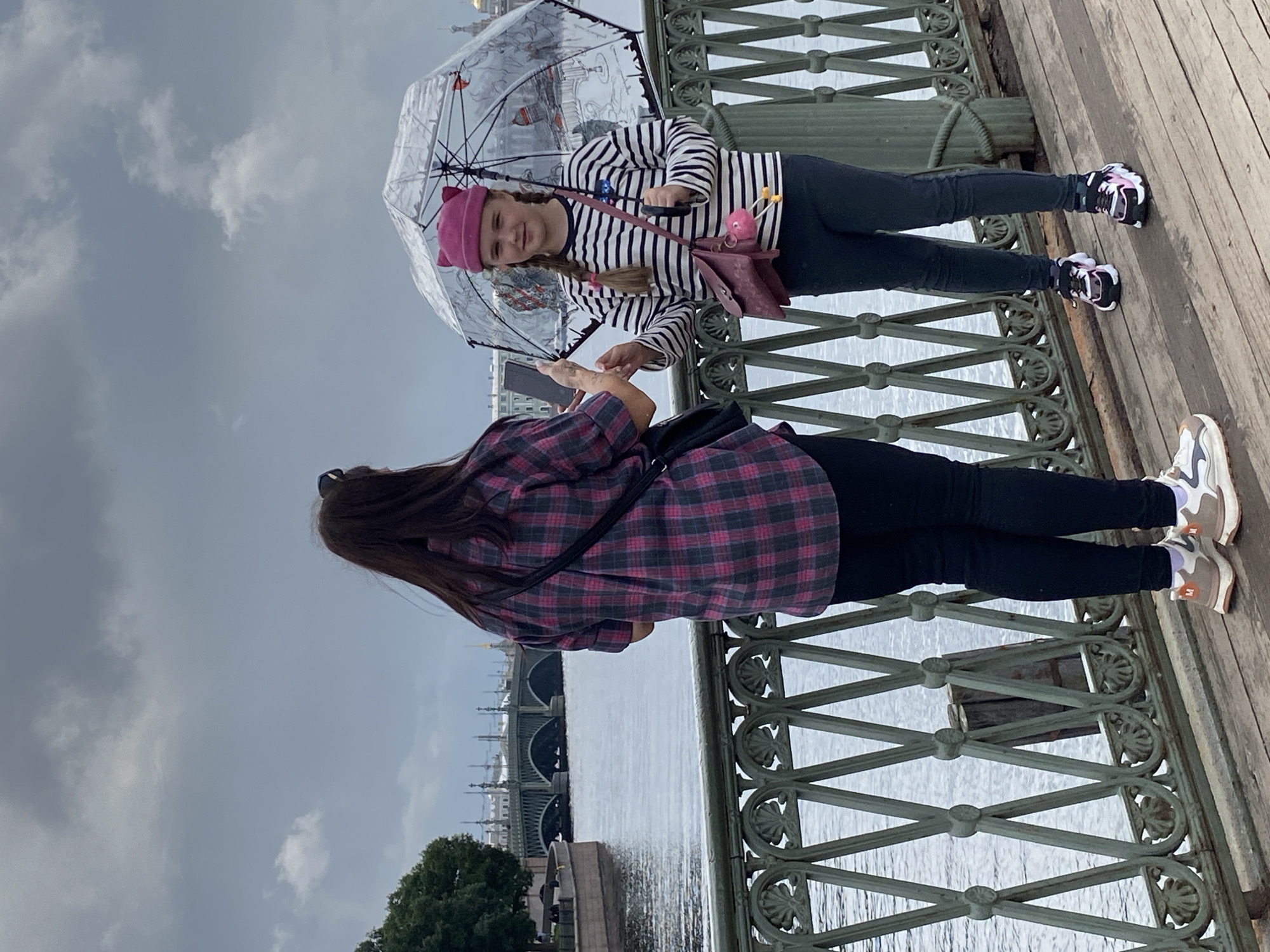 Two females are standing on a wooden pier with an ornate metal railing. A river with a bridge in the background is visible. The female on the left, wearing a plaid shirt, is holding a phone, possibly taking a picture. The female on the right, wearing a striped shirt and a pink hat, holds a clear umbrella with graphics. Both females are wearing dark pants and sneakers. The sky is overcast. The location is likely St. Petersburg, Russia, given the architectural style of the bridge and buildings in the background.