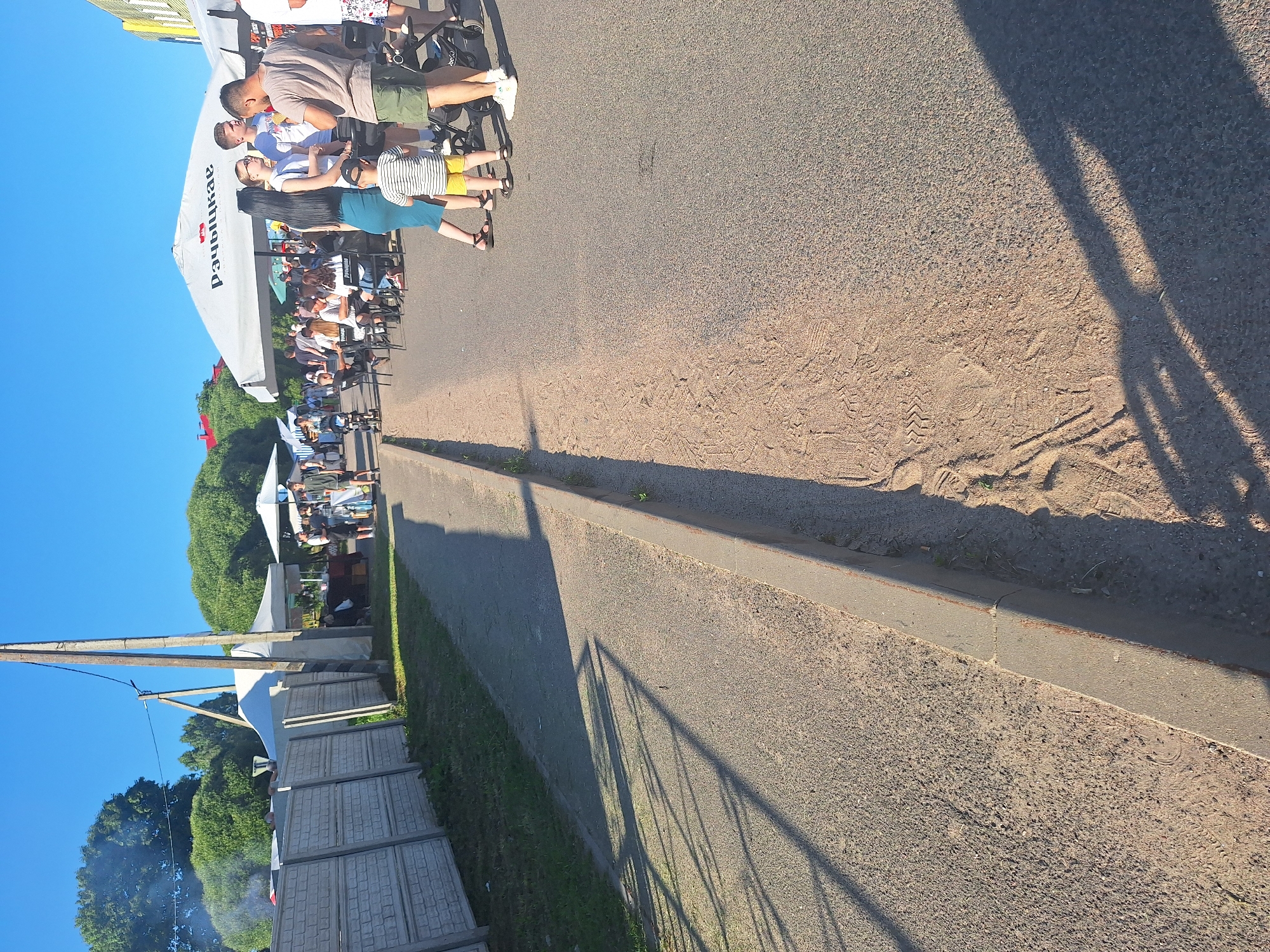 The image depicts an outdoor scene, likely a public event or market, under a clear blue sky. In the foreground, a paved area transitions to a sandy strip along a concrete curb. Further back, a group of people are gathered under large white umbrellas, suggesting an area for food or drink service. Some individuals are seated at tables, while others stand and converse. A young child in a striped shirt and yellow shorts is visible, interacting with adults. To the left, a fence made of concrete blocks runs along a grassy area, with a white tent or canopy visible behind it. Smoke emanates from a barbecue area behind the fence, indicating food preparation. A wooden utility pole with wires stands prominently on the left side of the image, casting long shadows across the pavement due to the sun's angle. The overall atmosphere suggests a warm, sunny day at an outdoor gathering. The location is specified as Блеўчыцкі сельскі Савет, Belarus.