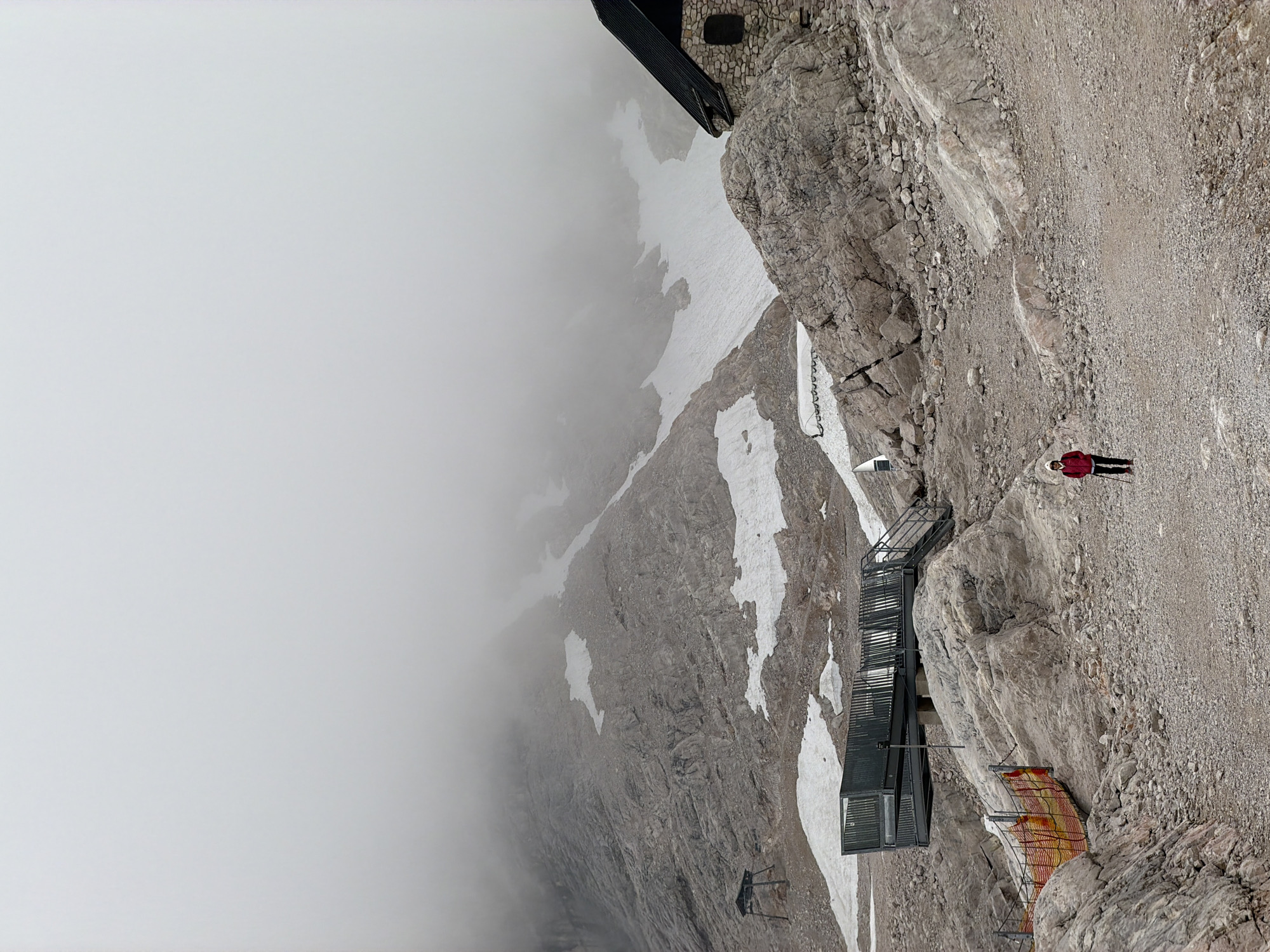 A lone hiker, dressed in a red jacket and white hat, stands on a gravel path in a rocky, mountainous terrain, holding a walking stick. The scene is set in Garmisch-Partenkirchen, Germany, likely at a high altitude due to the presence of snow patches on the mountainside and a cloudy, overcast sky. In the background, a rugged, rocky mountain face is partially obscured by fog, with remnants of snow clinging to its slopes. To the right, a stone building with a dark, sloped roof and a small window is visible. To the left, a metal viewing platform with stairs is constructed on stilts, overlooking the rocky landscape. A safety fence made of orange netting and metal poles borders a path in the foreground. The environment appears barren and rugged, with scattered rocks and gravel covering the ground.