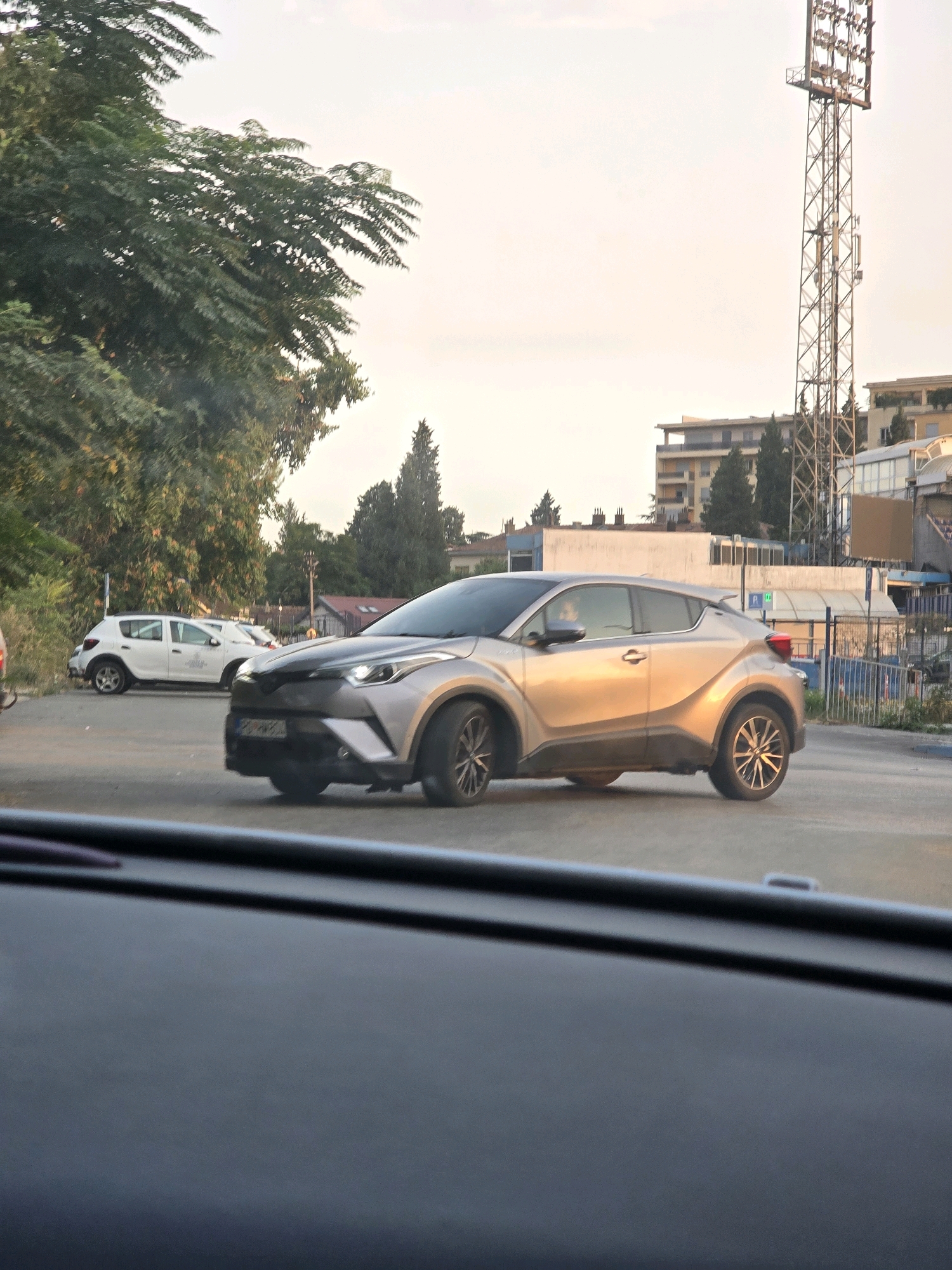 A silver Toyota C-HR is driving in an outdoor area. The car is the primary subject, positioned in the center of the frame. The vehicle has a sleek, modern design with distinctive headlights and taillights. The driver's side window is slightly open, and a person's head can be seen inside.
In the background, a white Dacia Sandero is parked to the left of the Toyota. Behind the cars, there are trees and buildings, suggesting an urban or suburban setting. A tall transmission tower with multiple platforms and lights is visible on the right side of the image, a common sight near sports stadiums. The sky is overcast with a pale, hazy quality, indicating either early morning or late afternoon light.
The scene appears to be a street or parking lot, possibly near a stadium, given the presence of the tower. There are no other people clearly visible in the image besides the driver. The overall atmosphere is ordinary and functional, a snapshot of daily life in Podgorica, Montenegro.