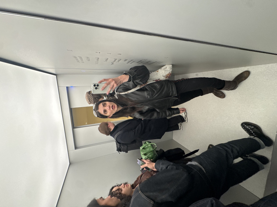 A group of people are in an elevator. A woman holds up a phone, presumably taking a photo in the mirror. She wears a black jacket, dark pants, and boots. To her side is a man in a black coat. Other people are visible in the reflection of the mirror, including someone using a phone and another wearing jeans and black shoes. The elevator has white walls, and writing is faintly visible on one wall.