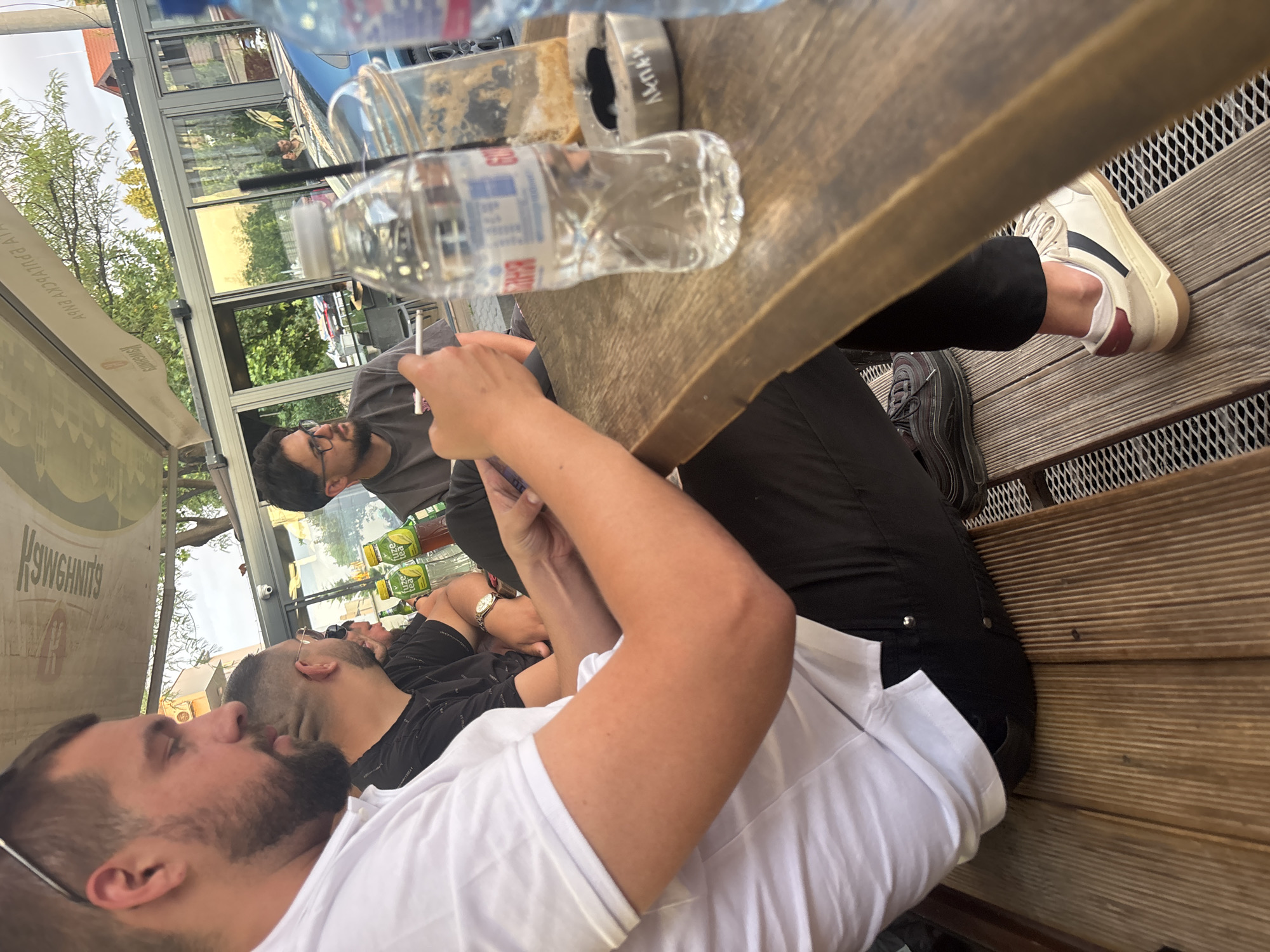 A medium, eye-level shot captures a group of three males sitting at an outdoor table in Plovdiv, Bulgaria. The foreground features a man wearing a white T-shirt, his attention directed out of frame, his left hand holding a smartphone. Partially visible, the man next to him is wearing a black shirt and a wristwatch. A man in a gray shirt and glasses is looking down at the table. The outdoor table contains water bottles, a glass with a straw, and an ashtray. Below the table are the legs and feet of the man in black pants and shoes. The table sits on a wooden platform with metal grate beneath. The background reveals an awning with lettering that says "KUSHWN'S", a view of a glass-enclosed restaurant or patio, and foliage.