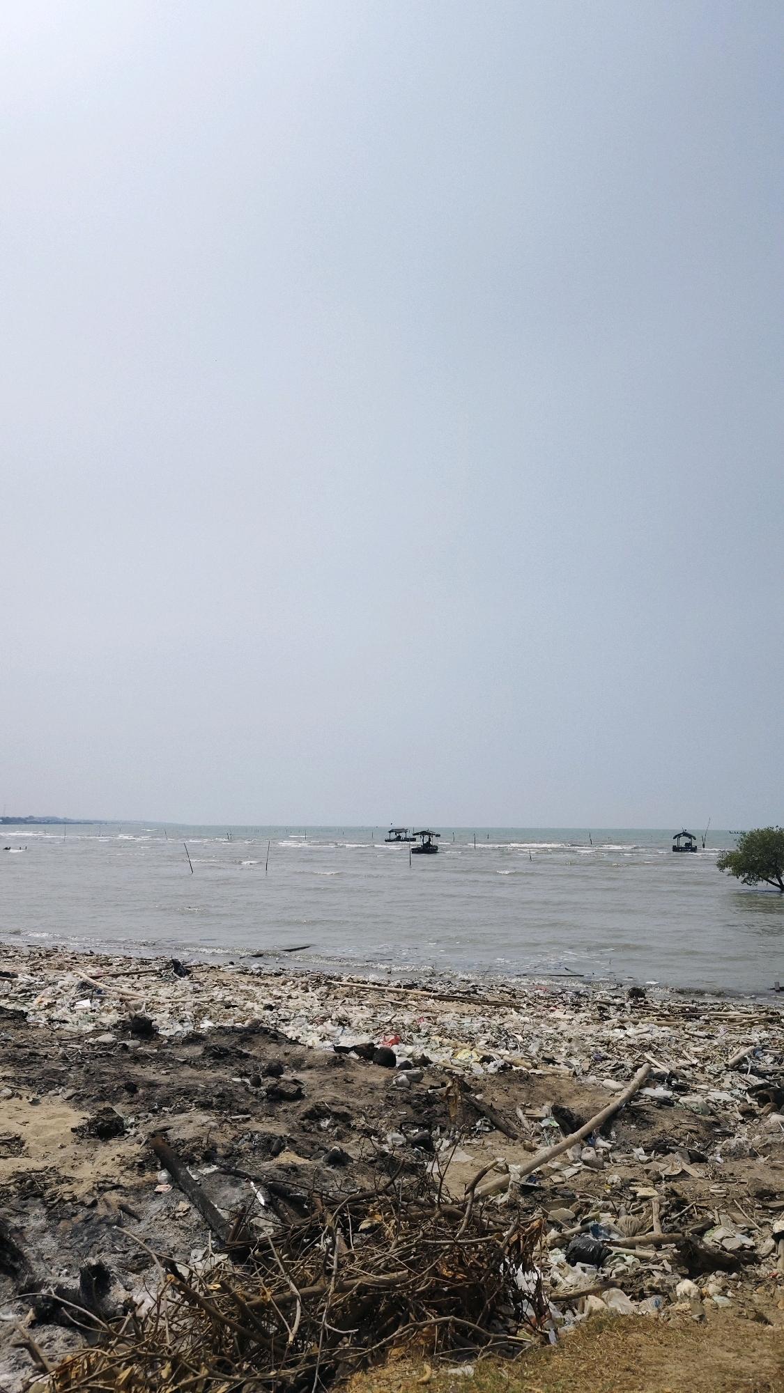 The image depicts a polluted beach in Pangpajung, Indonesia. The foreground is covered in a dense layer of trash, including plastic bags, bottles, and debris from burnt organic matter and wood. The beach transitions into a body of water, likely the sea, which is calm with small waves. In the distance, three small boats with awnings are visible on the water, along with several wooden poles sticking out of the sea. A tree is also visible on the right side of the image, leaning towards the water. The sky is overcast and pale. The overall scene conveys a sense of environmental neglect and pollution.