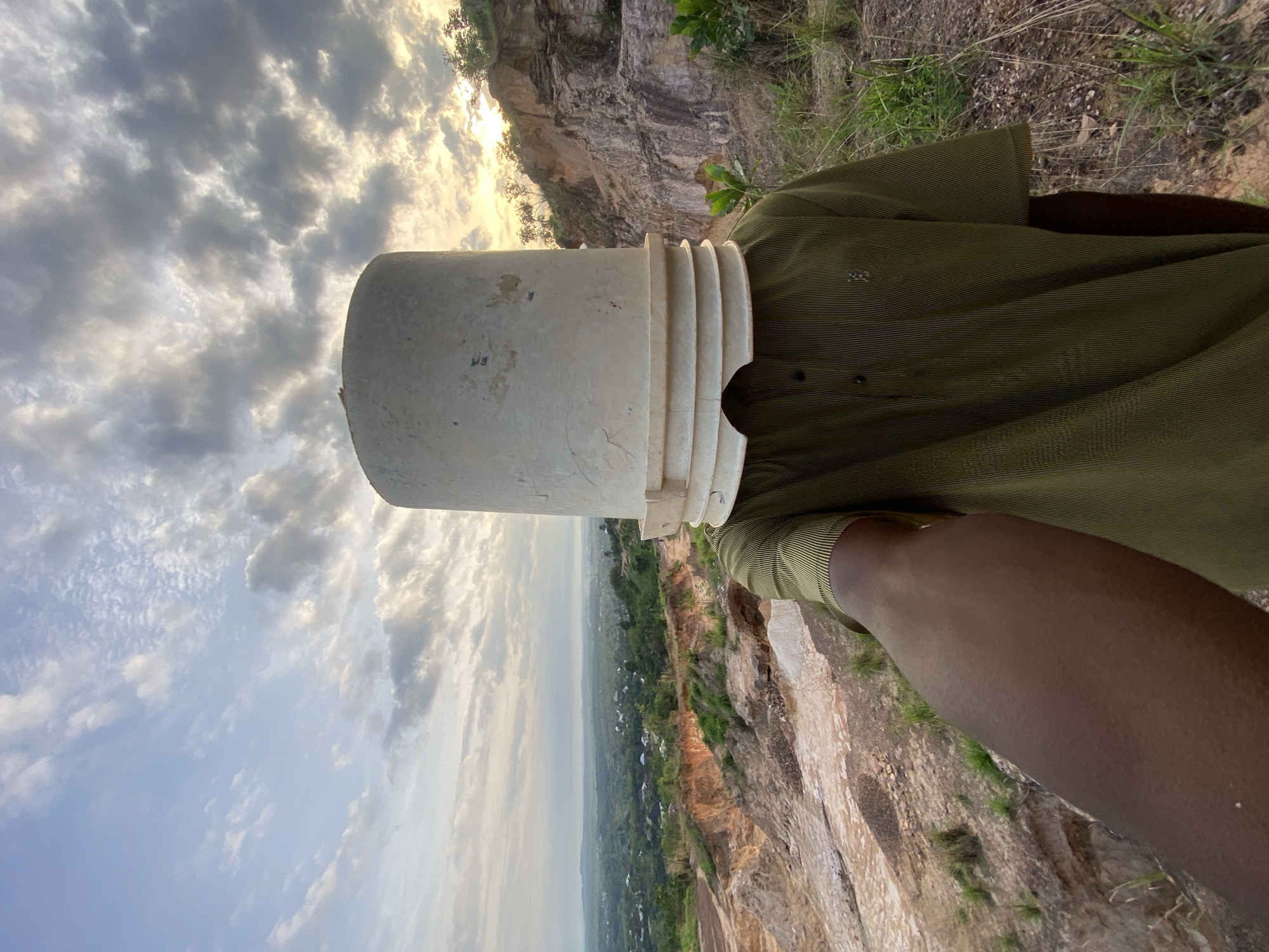 A person stands on a rocky cliff edge in Ufueseni, Nigeria, looking out over a vast landscape. A white plastic bucket is playfully placed upside down over their head. The person is wearing a dark olive green, horizontally striped short-sleeved shirt, and their dark-skinned left arm is visible in the foreground. The bucket is plain white with distinct concentric ridges around its open rim, which rests on the person's head.
The setting reveals a dramatic natural environment. In the immediate vicinity of the person are rugged, exposed light-colored rock formations, suggesting a quarry or natural erosion. Sparse green grass and small bushes grow in patches on the rocky ground. Beyond the cliff, a sweeping vista unfolds, featuring a wide valley or plain covered in lush green vegetation. Numerous small structures, indicative of a village or scattered settlements, are visible in the distance, extending towards the hazy horizon where faint hills or mountains can be discerned.
The sky is partly cloudy, with a mix of grey and white clouds. A warm, golden light, emanating from the setting or rising sun, illuminates the clouds and the distant landscape, creating soft shadows and a serene atmosphere. The overall scene captures a moment of playful interaction with a grand natural backdrop in West Africa.