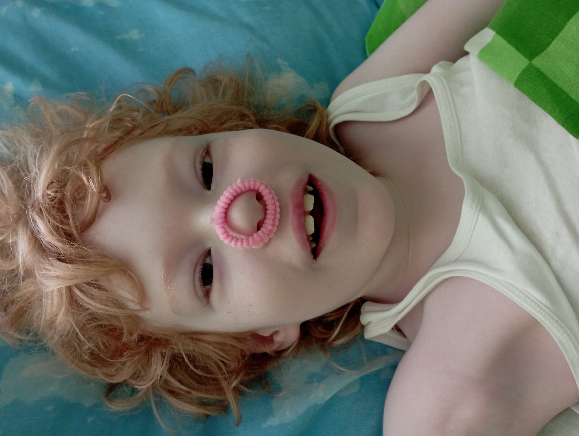 A close-up, overhead shot shows a young person with ginger hair and freckled skin, lying on a turquoise-colored surface, possibly a bed. A pink hair tie is positioned on their nose. The person is wearing a white tank top, and a green and white checkered fabric is visible on the right side of the frame. The image appears to be taken indoors. There is no information in the image to suggest a specific location in Blagoveshchensk, Russia.