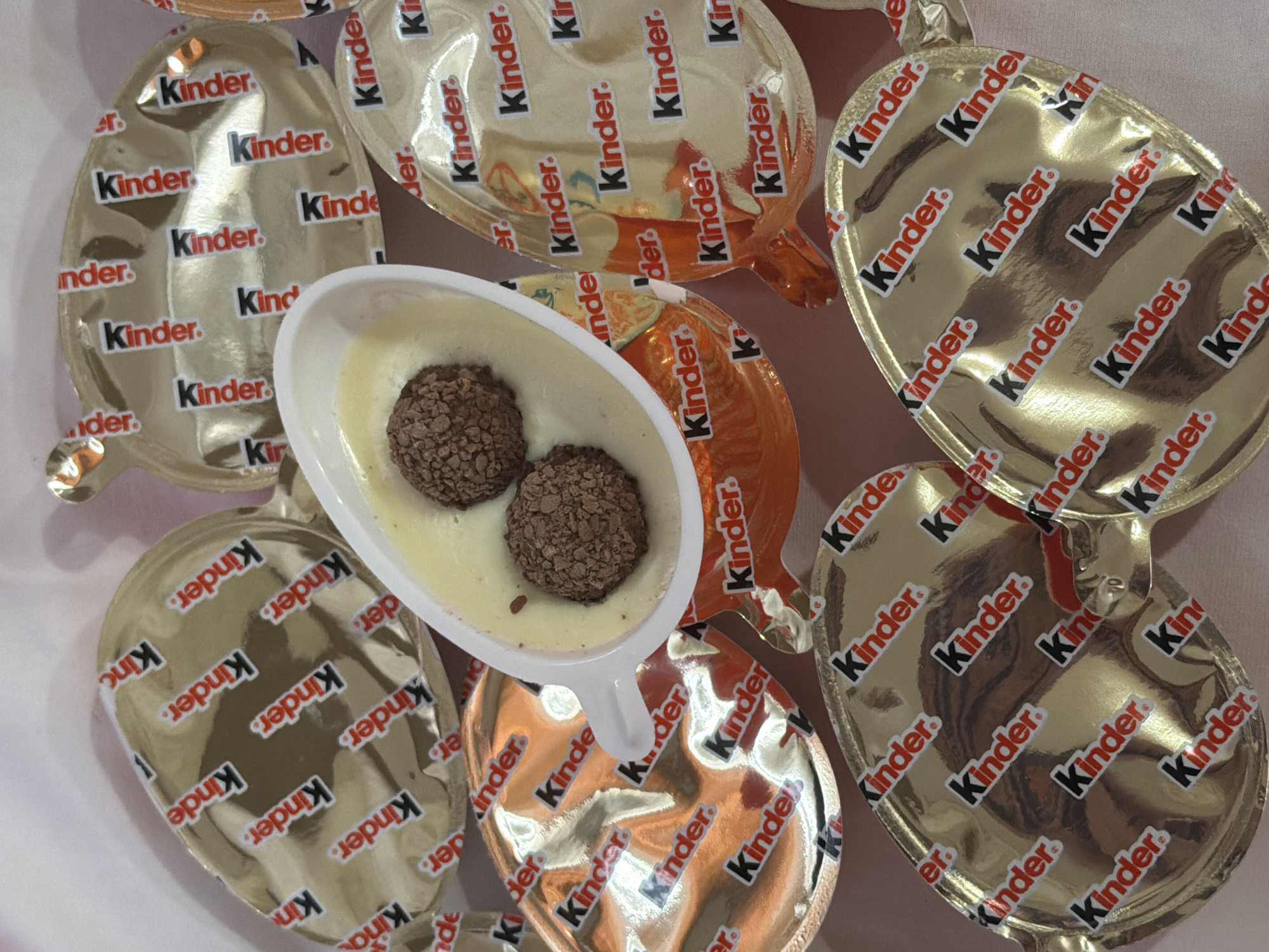 The image shows a collection of Kinder Joy chocolate eggs, both unopened and opened, on a light pink surface. The unopened eggs are individually wrapped in foil, with the "Kinder" logo repeated across the surface. In the center, a white plastic container, likely from an opened Kinder Joy egg, holds a white creamy substance with two round, chocolate-covered balls resting inside. The background and overall arrangement suggest a casual, possibly domestic setting.