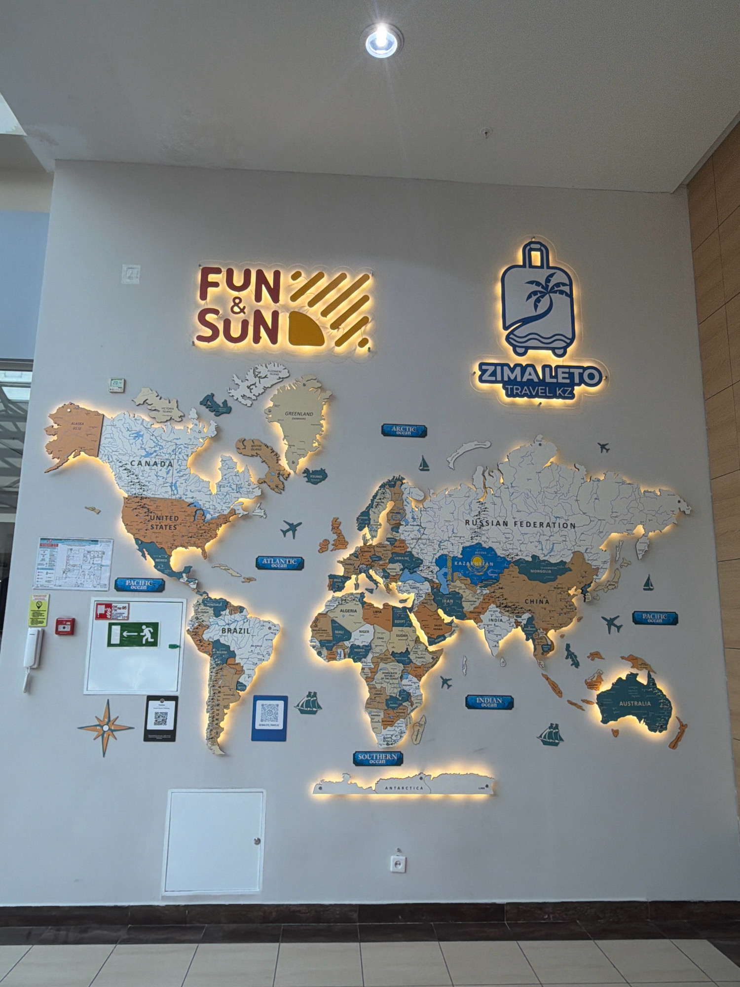 In Pavlodar, Kazakhstan, a large, illuminated world map is mounted on a light gray wall. The map is constructed from separate wooden pieces representing continents and countries, with country names printed in a dark color. Several bodies of water are labeled, including the Atlantic Ocean, Pacific Ocean, Indian Ocean, Arctic Ocean, and Southern Ocean.
Above the map, two neon signs are visible. The left sign displays "FUN & SUN" in red and yellow letters with yellow stripes and a sun graphic. The right sign features a blue silhouette of a suitcase with a palm tree and waves, along with the text "ZIMALETO TRAVEL KZ" in a blue oval.
Several smaller objects are also on the wall:
* To the left of the map, there's a white fire alarm panel, a white telephone, and a green and white exit sign with a running figure.
* A small digital display shows "20".
* A dark square with a QR code is placed to the left of Brazil.
* Another dark square with a QR code, labeled "SIMPLIFY", is located near Australia.
* A compass rose graphic is positioned to the left of Brazil.
* Several small airplane and sailboat icons are scattered across the map.
The wall is illuminated by ambient lighting from above and by the internal lights of the map and neon signs. The lower part of the image shows a dark brown skirting board and a floor with light and dark square tiles.